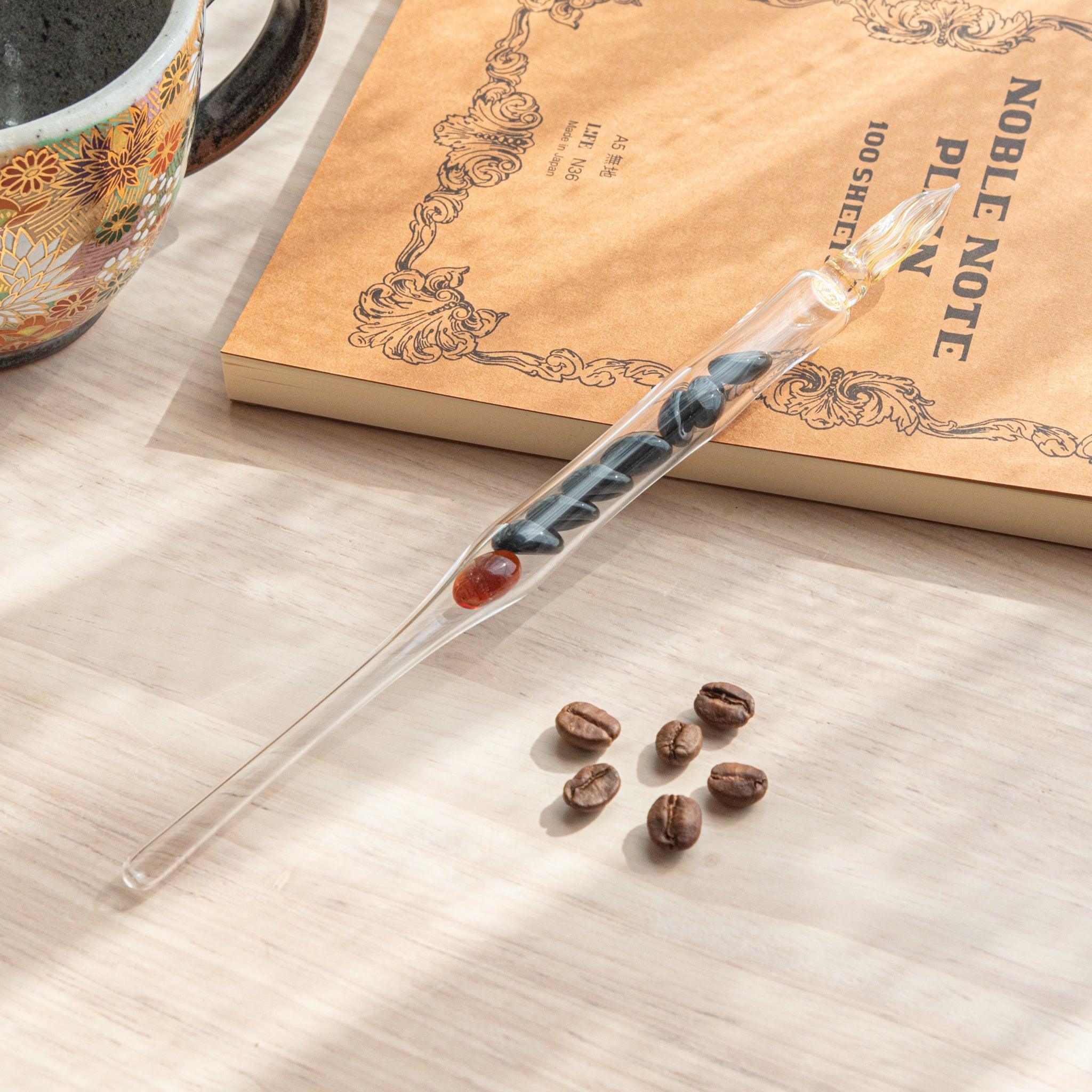 A glass dip pen with coffee bean-shaped glass pieces inside its shaft and a coffee-stained colored nib rests on a wooden surface near coffee beans and a plain notebook.