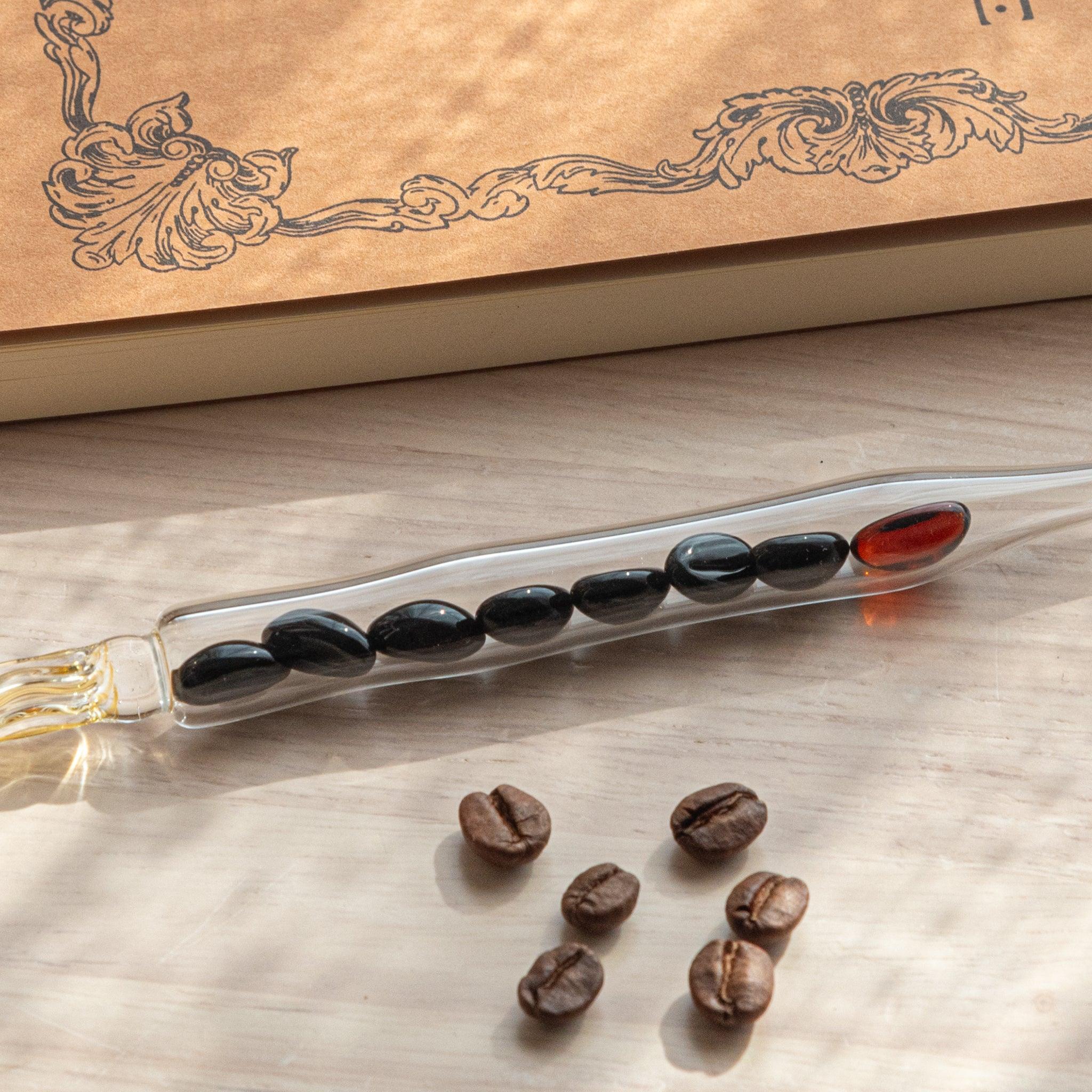 A glass dip pen with coffee bean-shaped glass pieces inside its shaft and a coffee-stained colored nib rests on a wooden surface near coffee beans and a plain notebook.