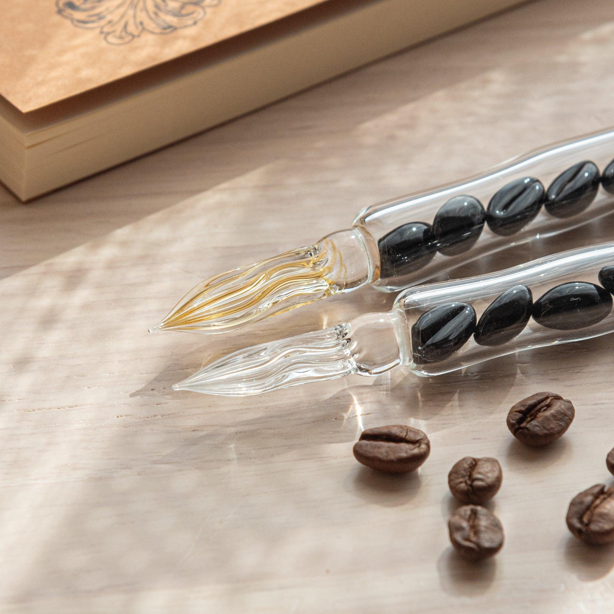 Two glass dip pens with coffee bean-shaped glass pieces inside their shafts are placed on a wooden surface near coffee beans and a plain notebook.