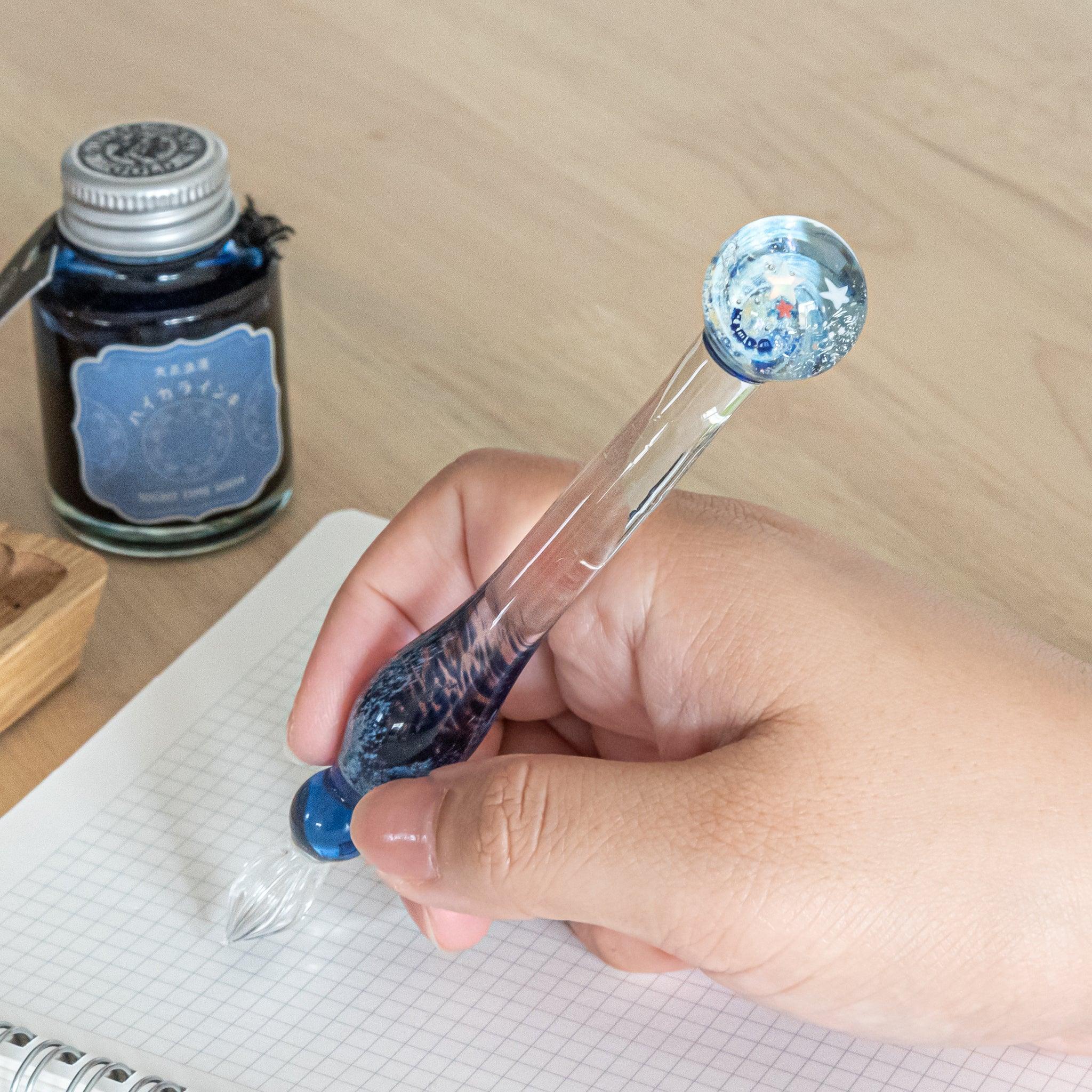 Hand holding a glass pen with a star-decorated top, writing on a notebook. Ink bottle labeled "Night Tree Moon" and other items are placed on the desk.