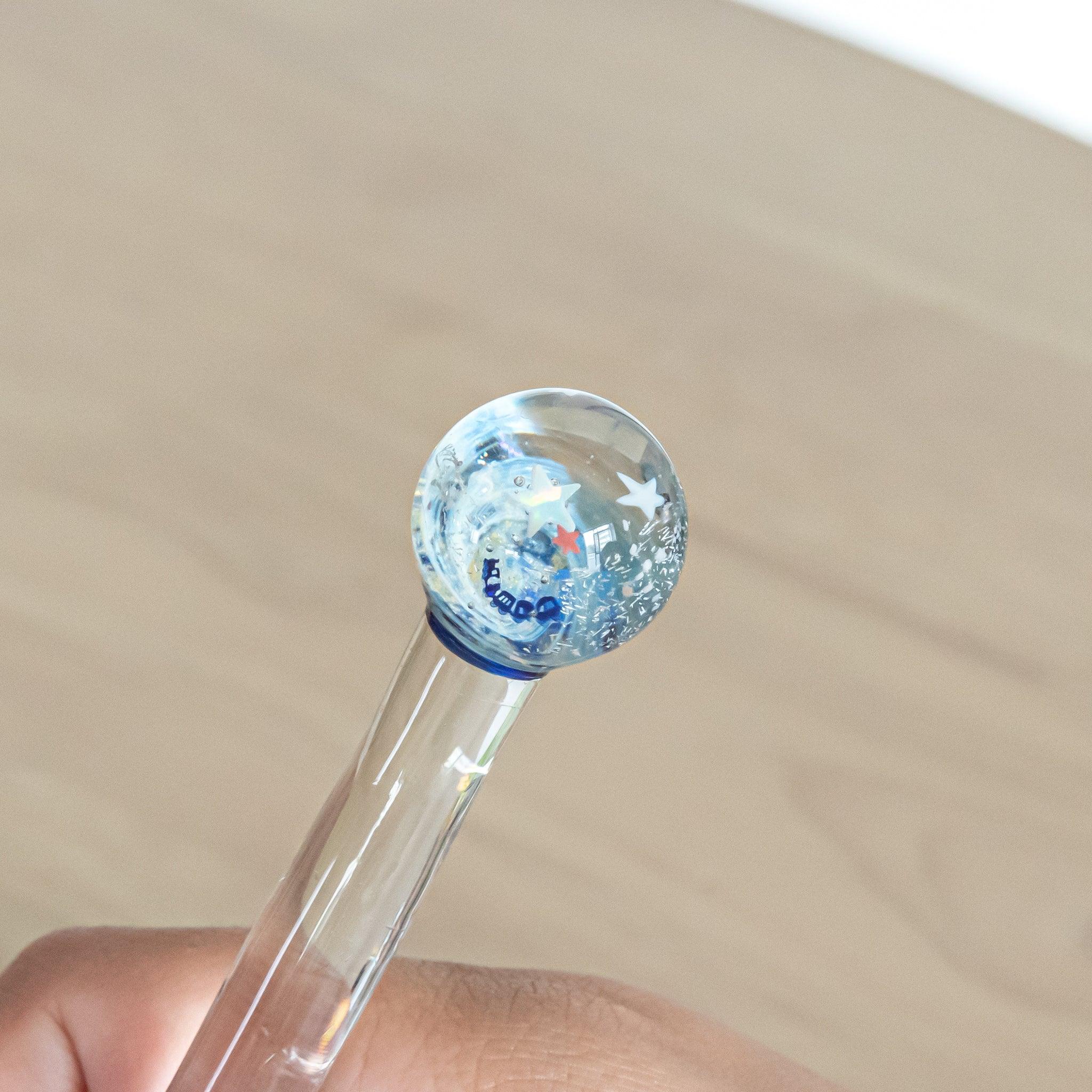 Close-up of a glass pen’s top with star designs, held in hand. The transparent shaft has a blue tint, with a blurred background.