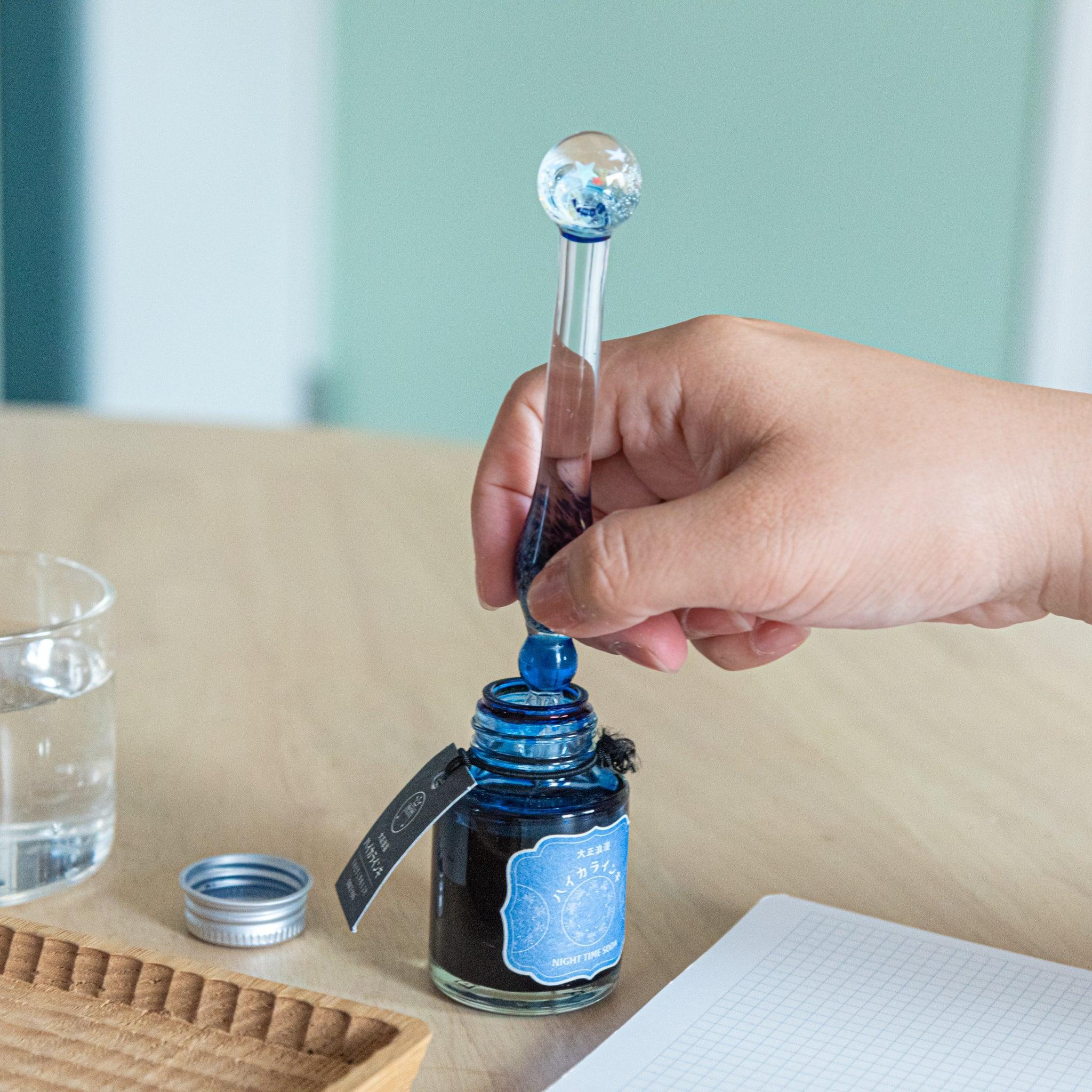 Hand dipping a star-topped glass pen into a blue ink bottle labeled "Night Tree Moon." Notebook and small items are arranged on the desk.