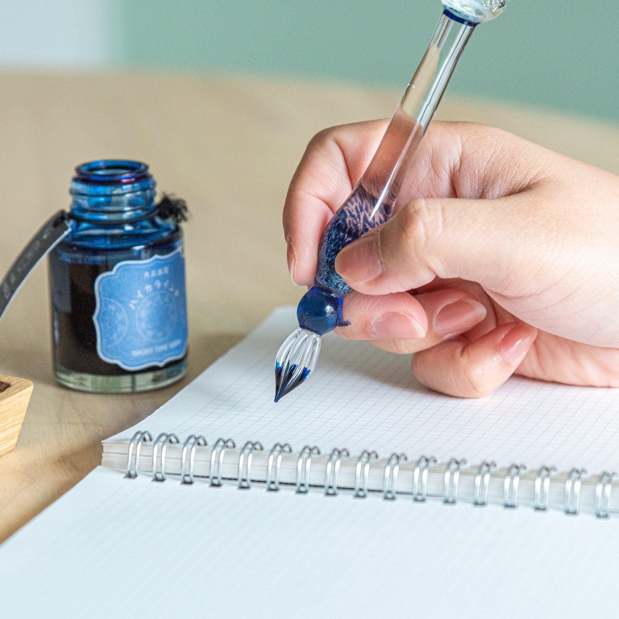 Hand holding a glass dip pen with a blue-tinted design, poised to write on a grid notebook. An open ink bottle labeled "Night Tree Moon" is nearby.