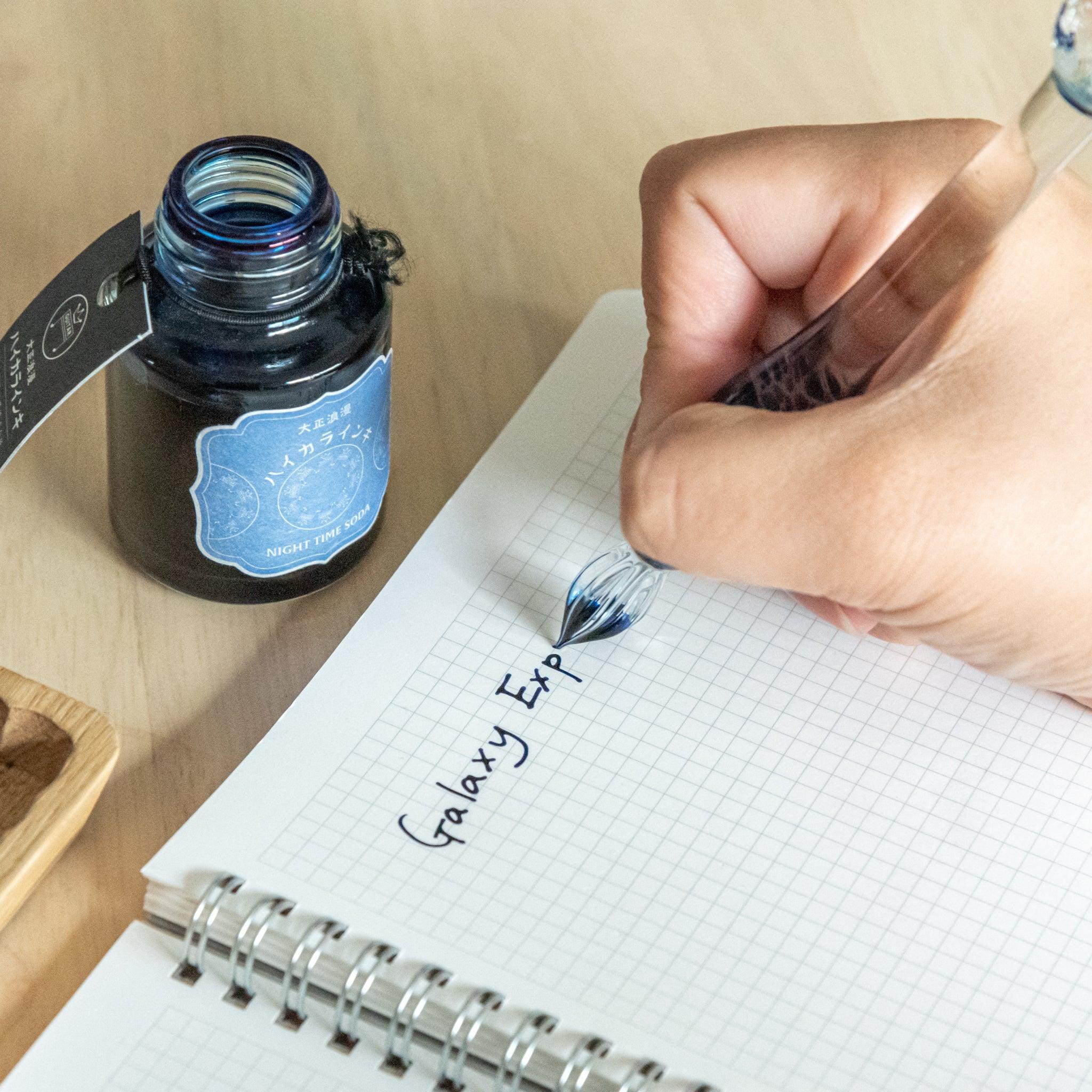 Hand writing "Galaxy Exp" on a grid notebook with a glass dip pen, next to an open ink bottle labeled "Night Tree Moon" on a wooden desk.