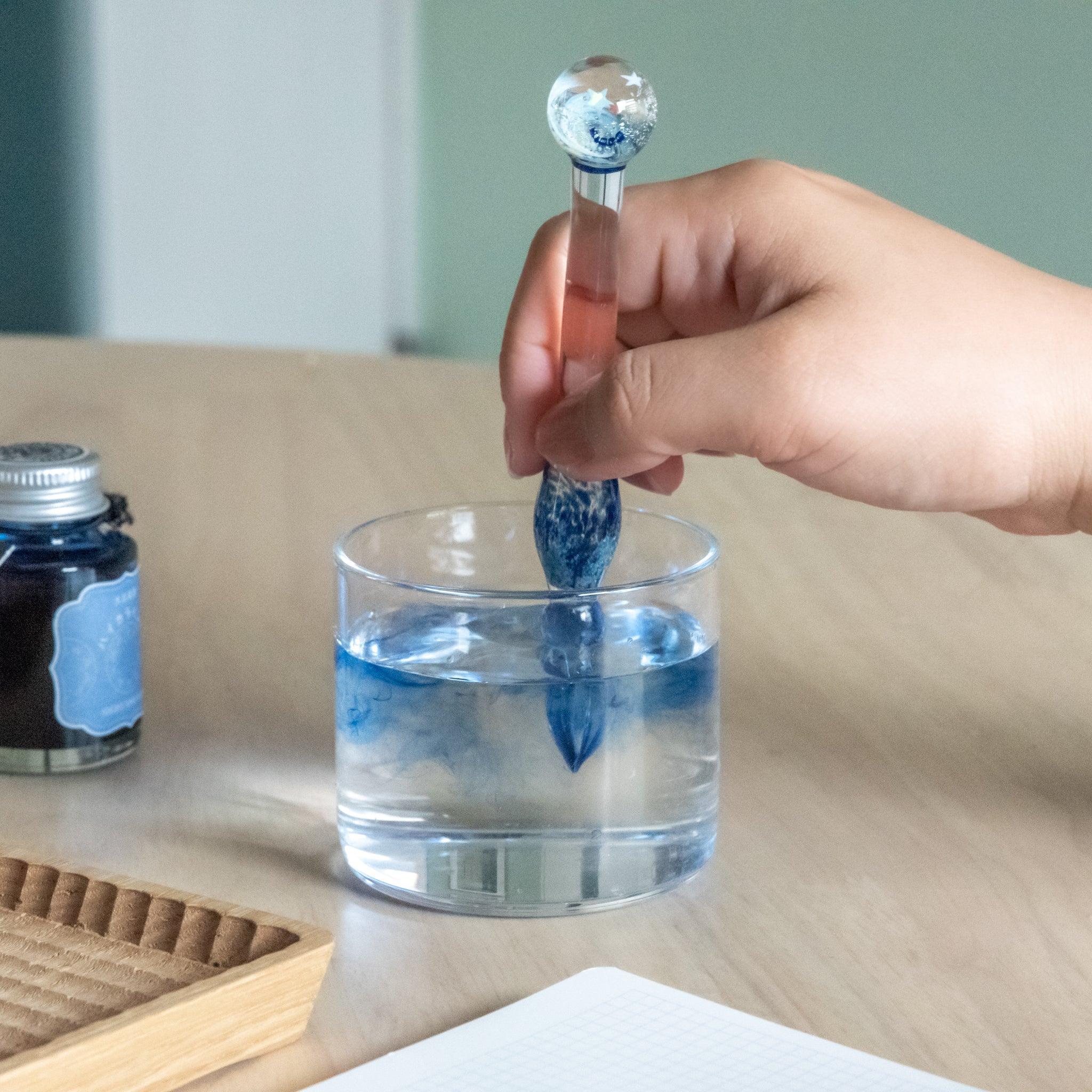 Person cleaning a star-topped glass dip pen in a clear glass of water, with blue ink swirling inside. Ink bottle and wooden tray on desk.