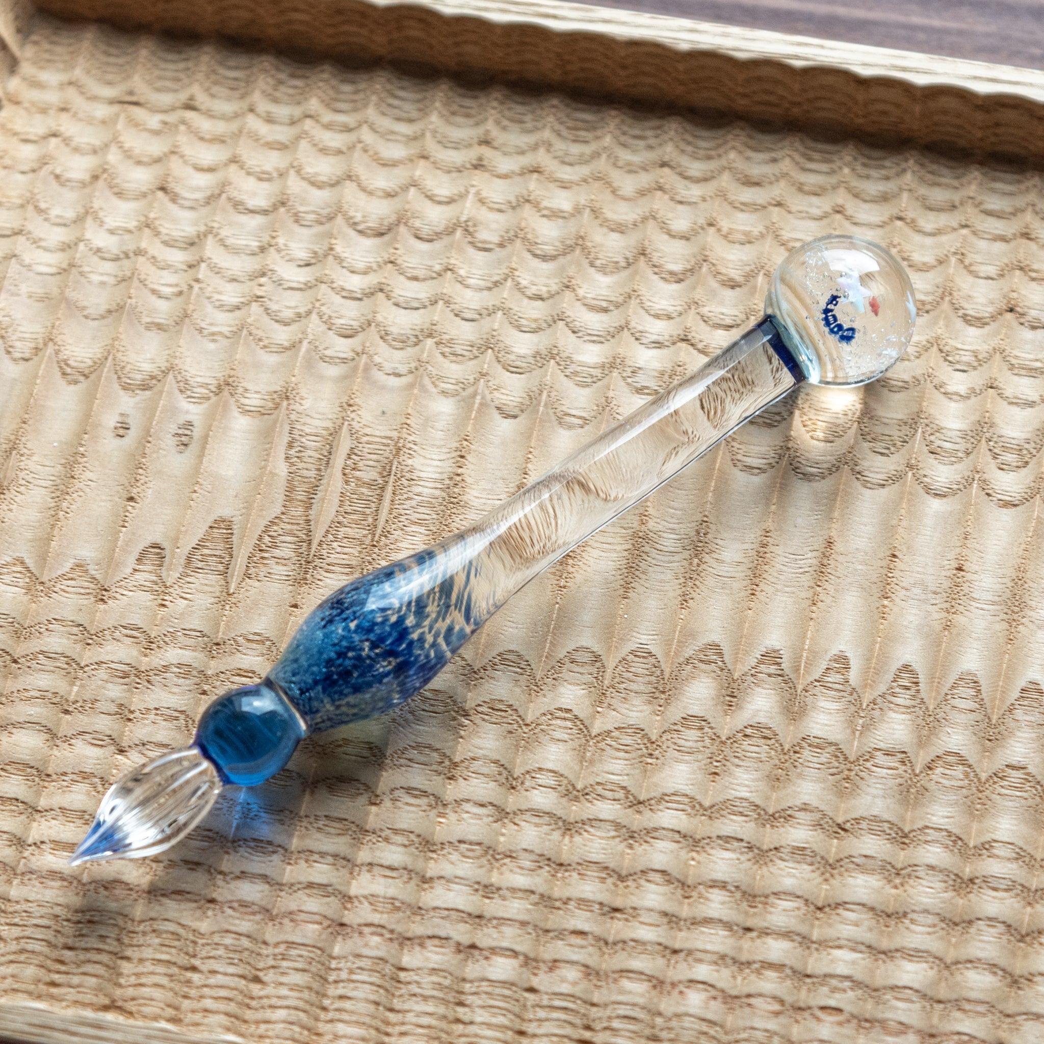 A decorative glass dip pen with a blue and clear design, featuring a star-decorated top, placed on a textured wooden tray.