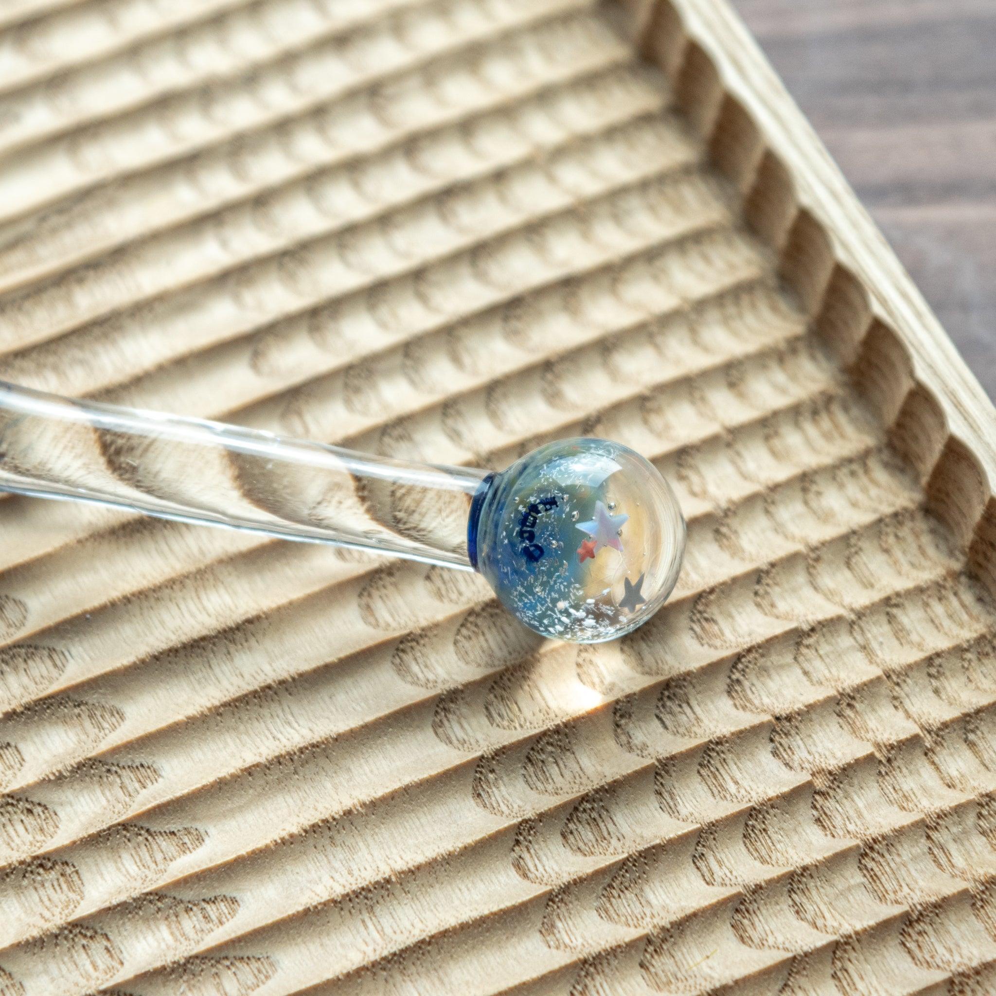 Glass dip pen with a star-decorated spherical top, resting on a textured wooden tray with a natural, wavy pattern.