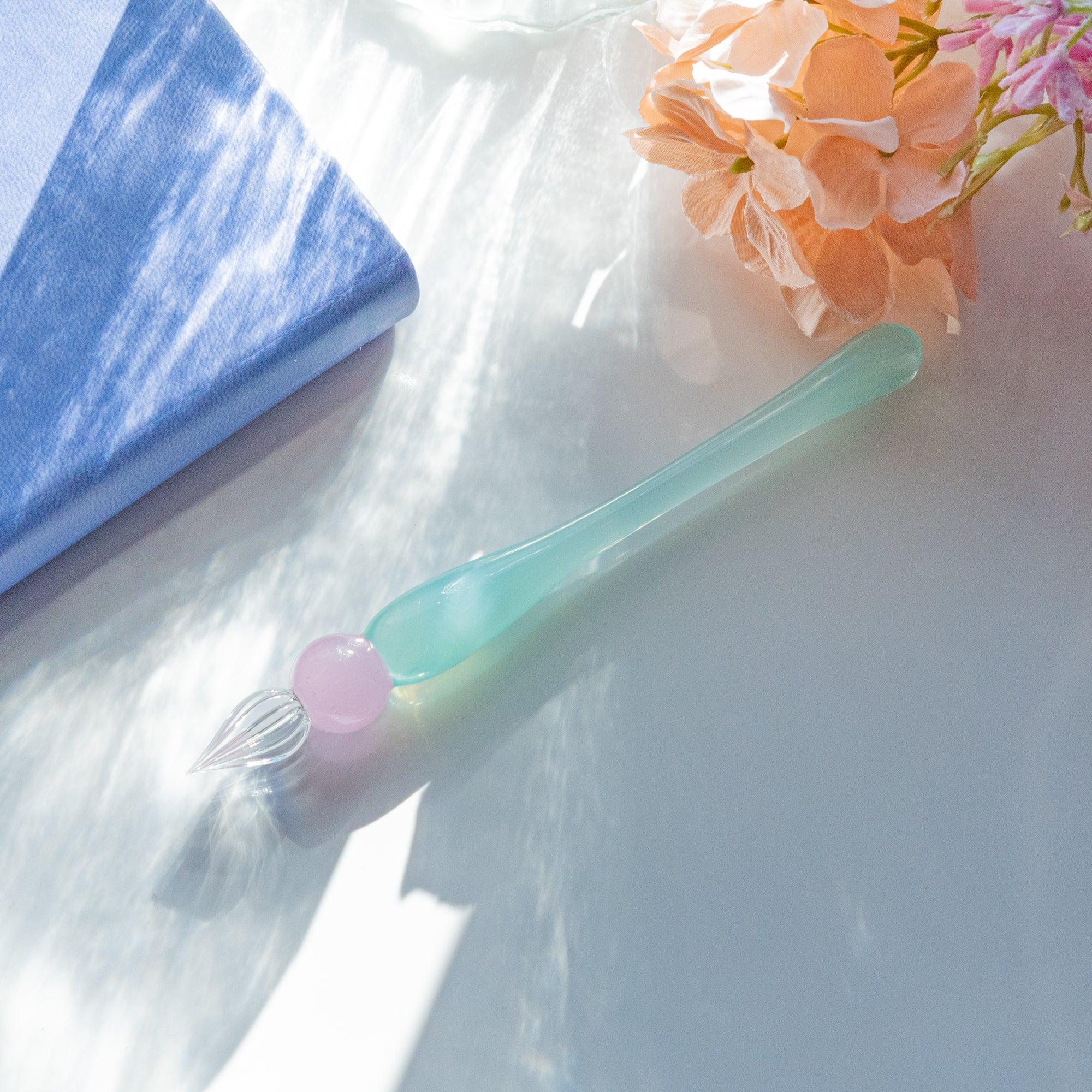 Pastel glass dip pen with a pink and green gradient, placed on a sunlit surface next to a blue notebook and soft pink flowers.