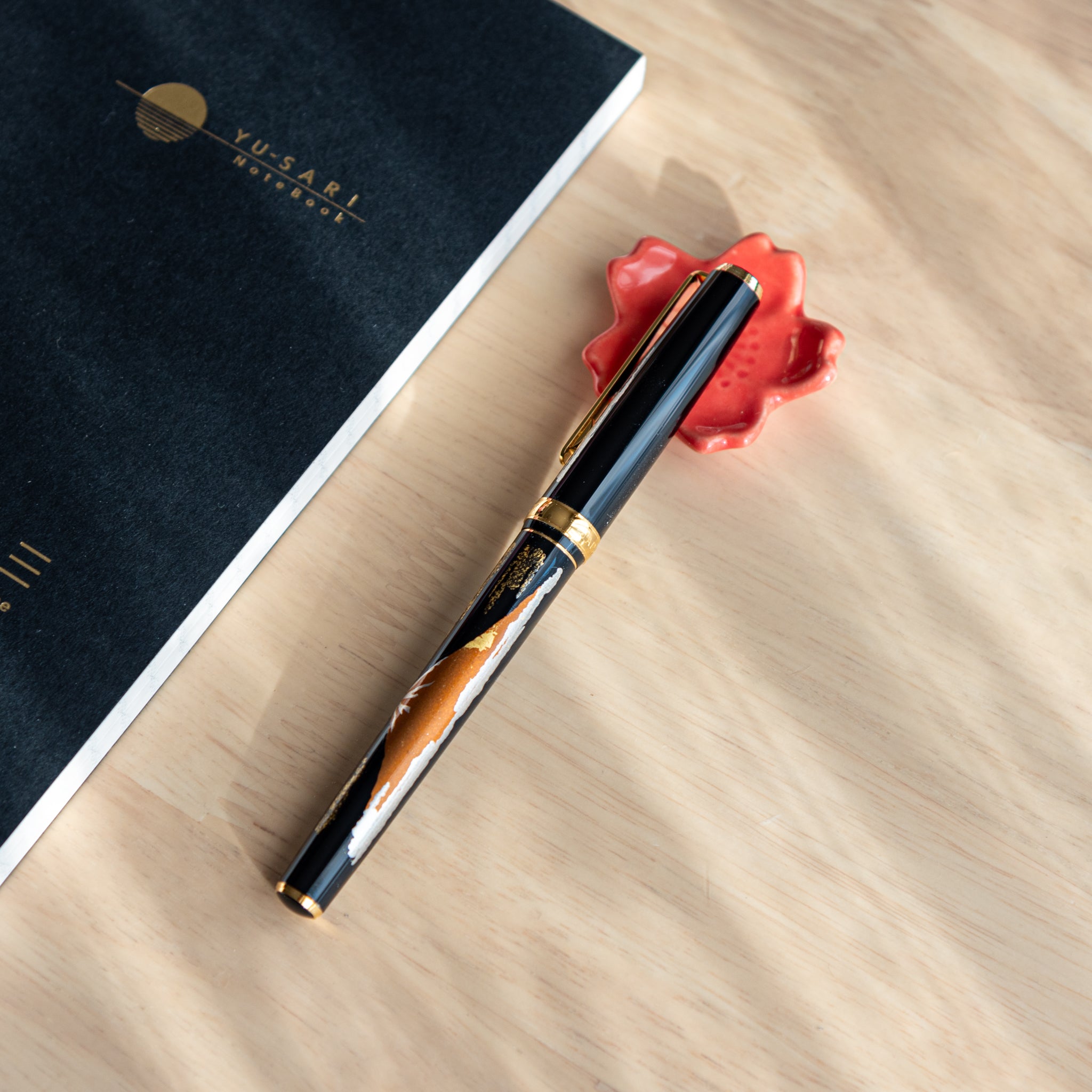 A Japanese fountain pen with gold and silver maki-e design rests on a red maple leaf-shaped holder. A black Yusari notebook lies nearby on a wooden desk.  