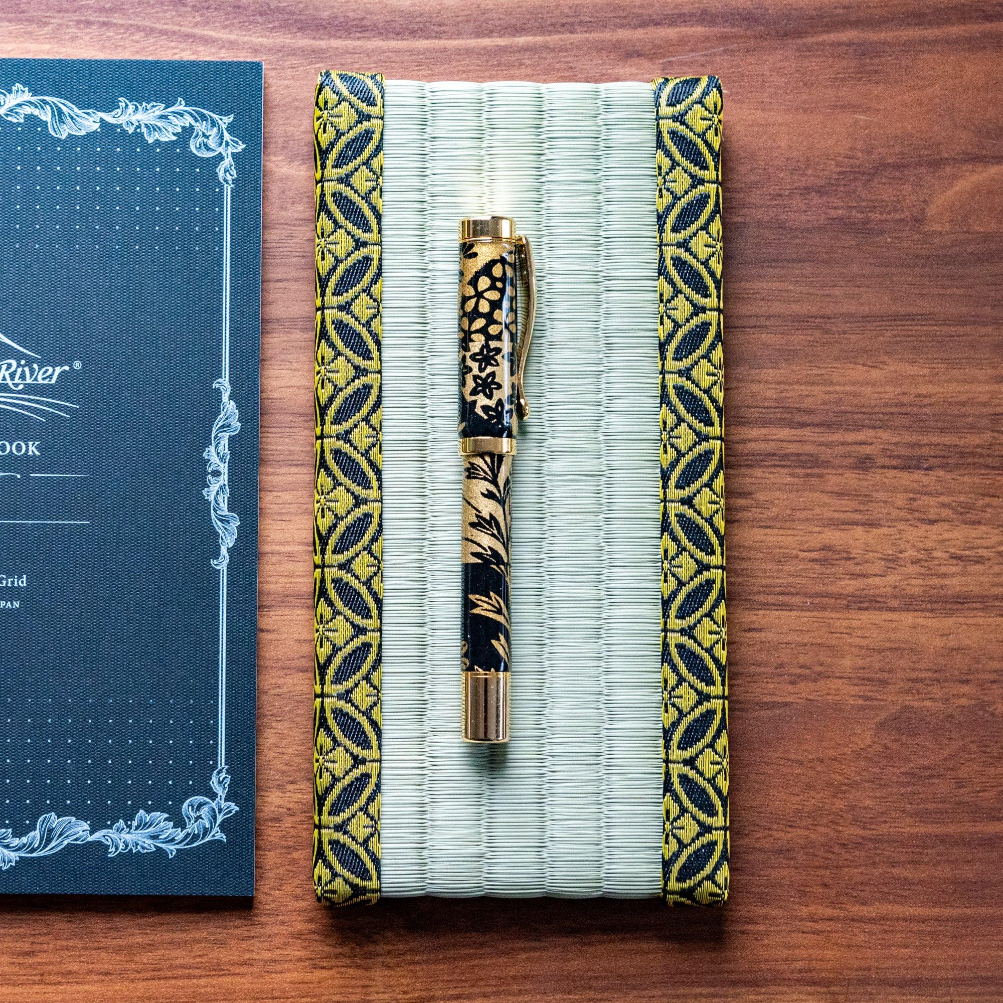 Japanese tatami pen tray with gold-patterned borders, paired with a black and gold pen, displayed beside a Tomoe River notebook on a wooden surface.  