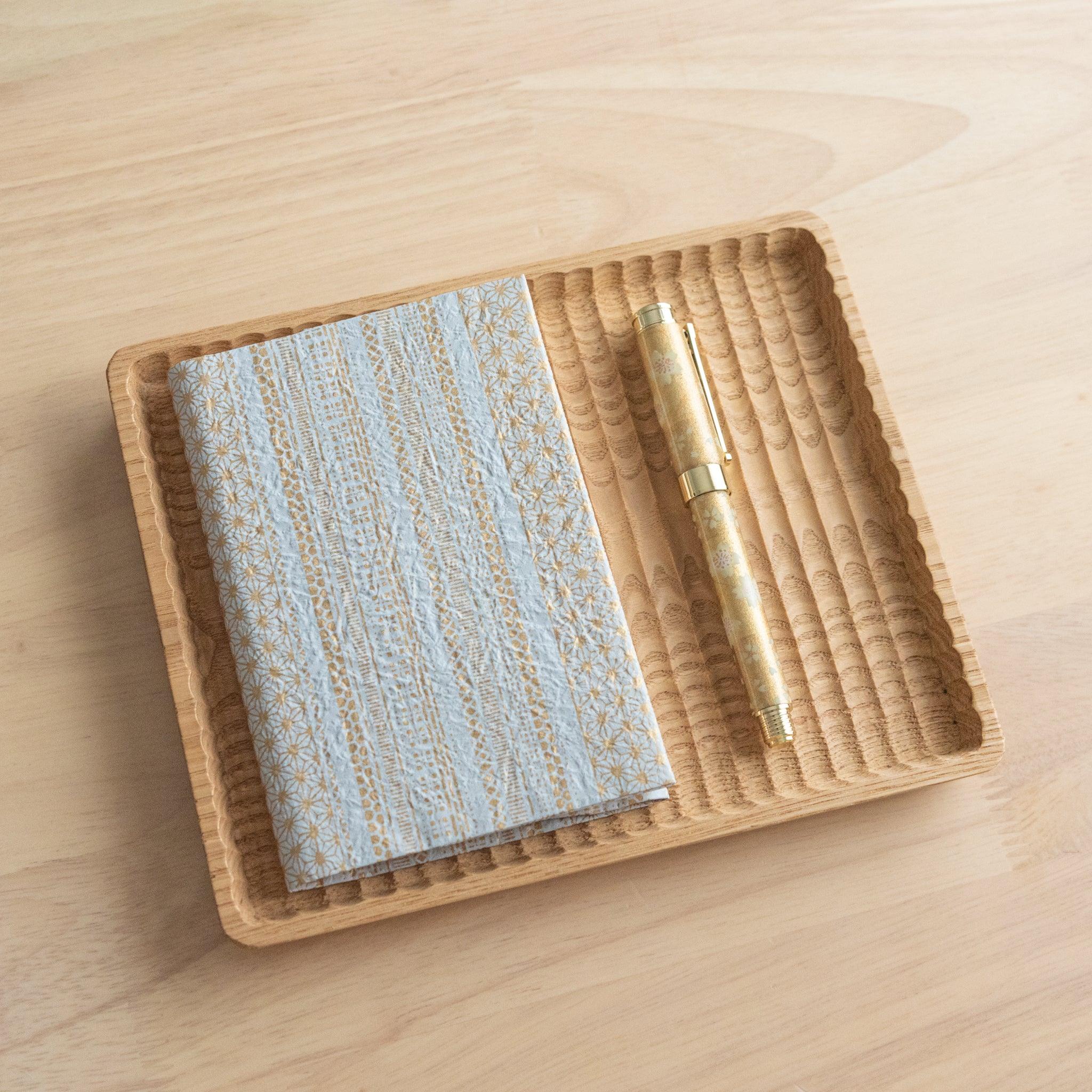 A blue and gold patterned notebook and a matching gold pen arranged on a wooden tray, placed on a light wooden surface.