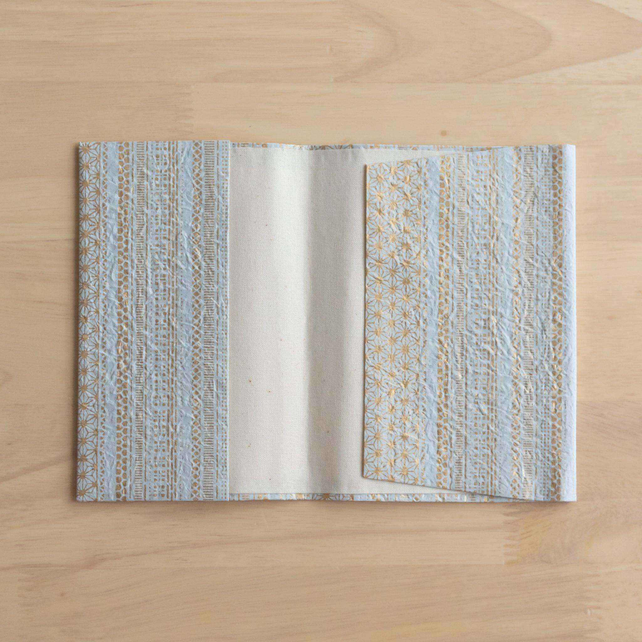 An open notebook cover with a blue and gold striped pattern, revealing its inner white lining on a light wooden surface.