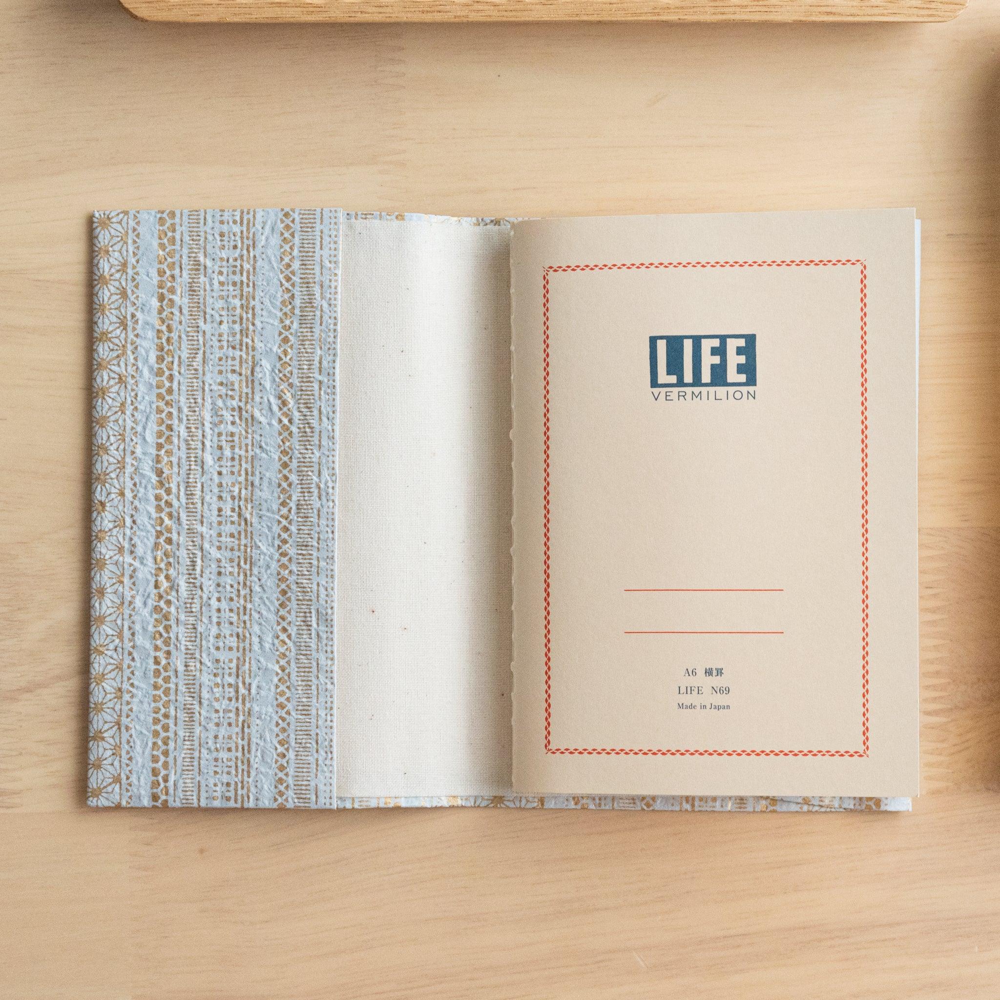 An open notebook with a blue and gold patterned cover, revealing a title page labeled "Life Vermilion" on a wooden surface.