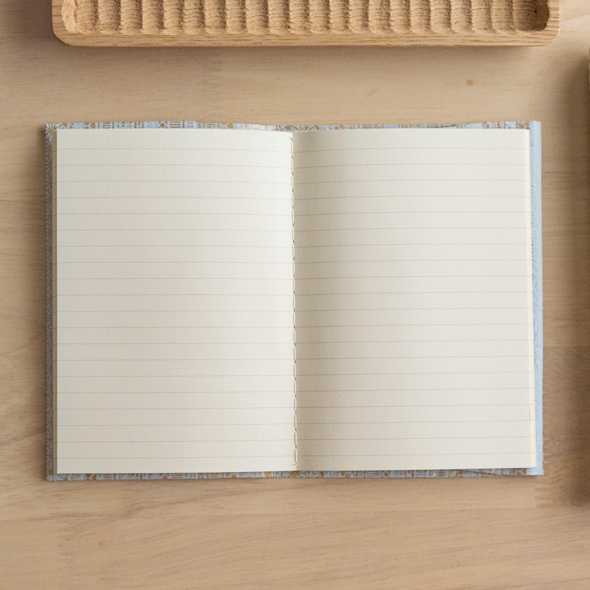 An open notebook with blue and gold patterned covers, fully revealing two lined pages, placed on a light wooden surface.