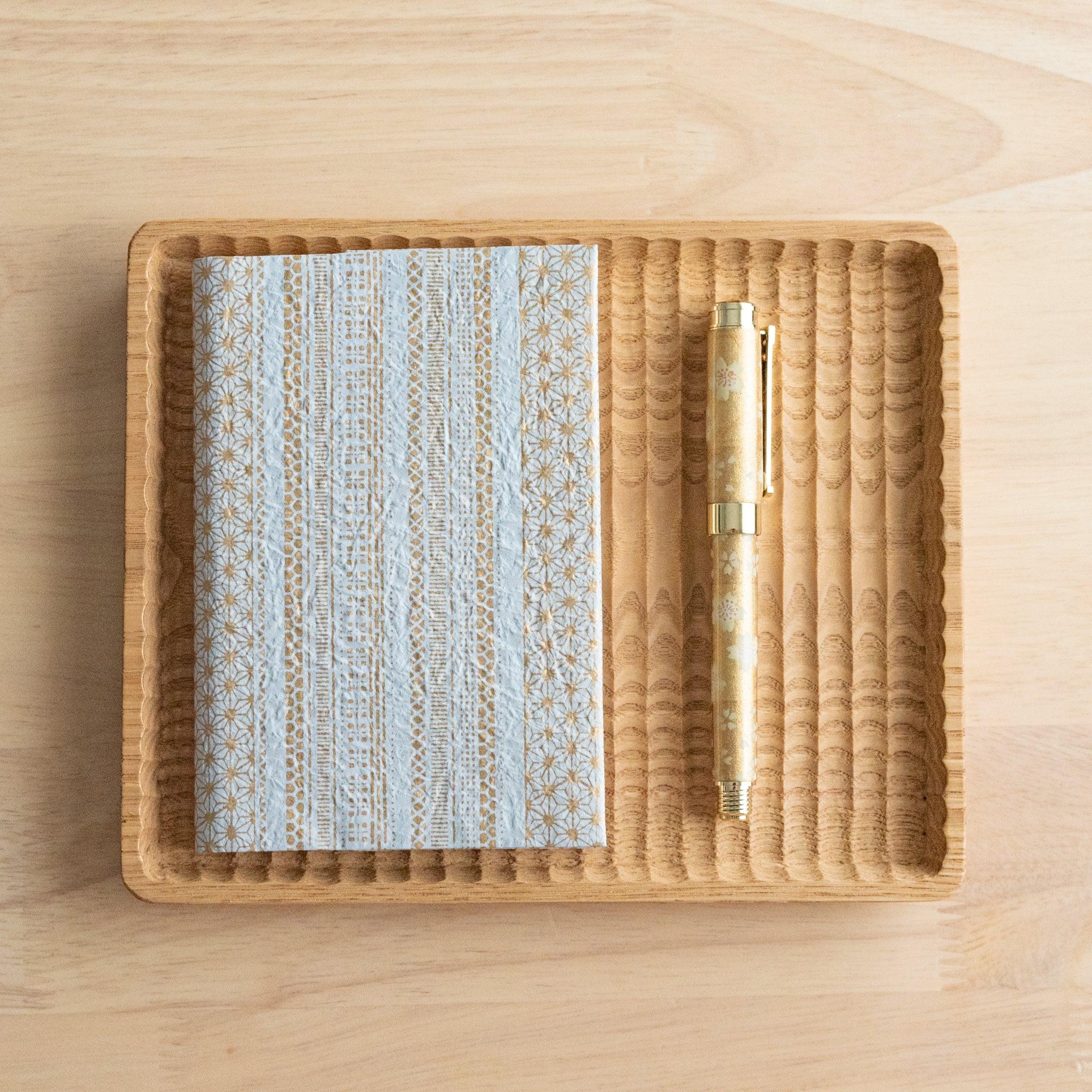 A notebook with a blue and gold striped pattern and a matching gold pen, placed neatly on a wooden tray on a light wooden surface.