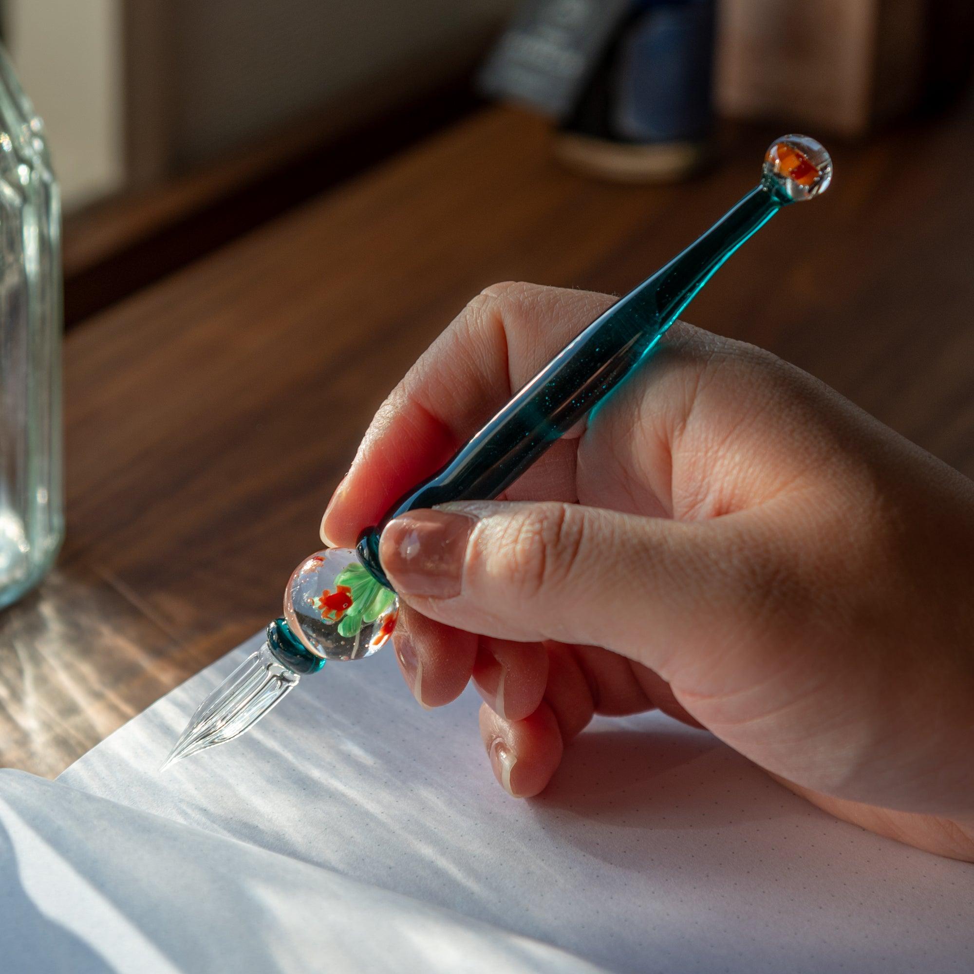 Hand holding a glass pen with goldfish motif inside, resembling a fishbowl, ready to write.