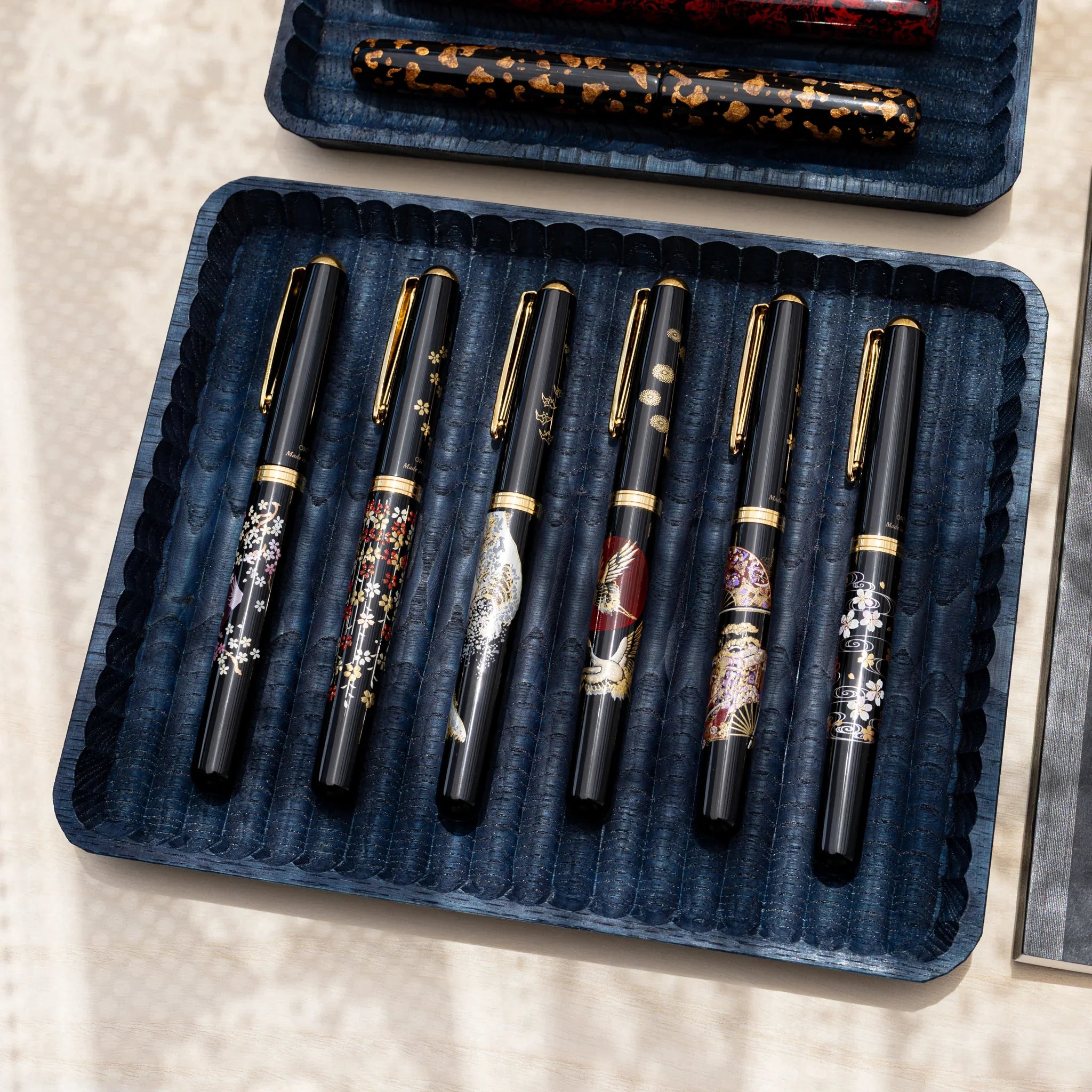 Indigo dyed wide and slim chestnut pen trays on a pale wood desk, the wide tray holding six black fountain pens