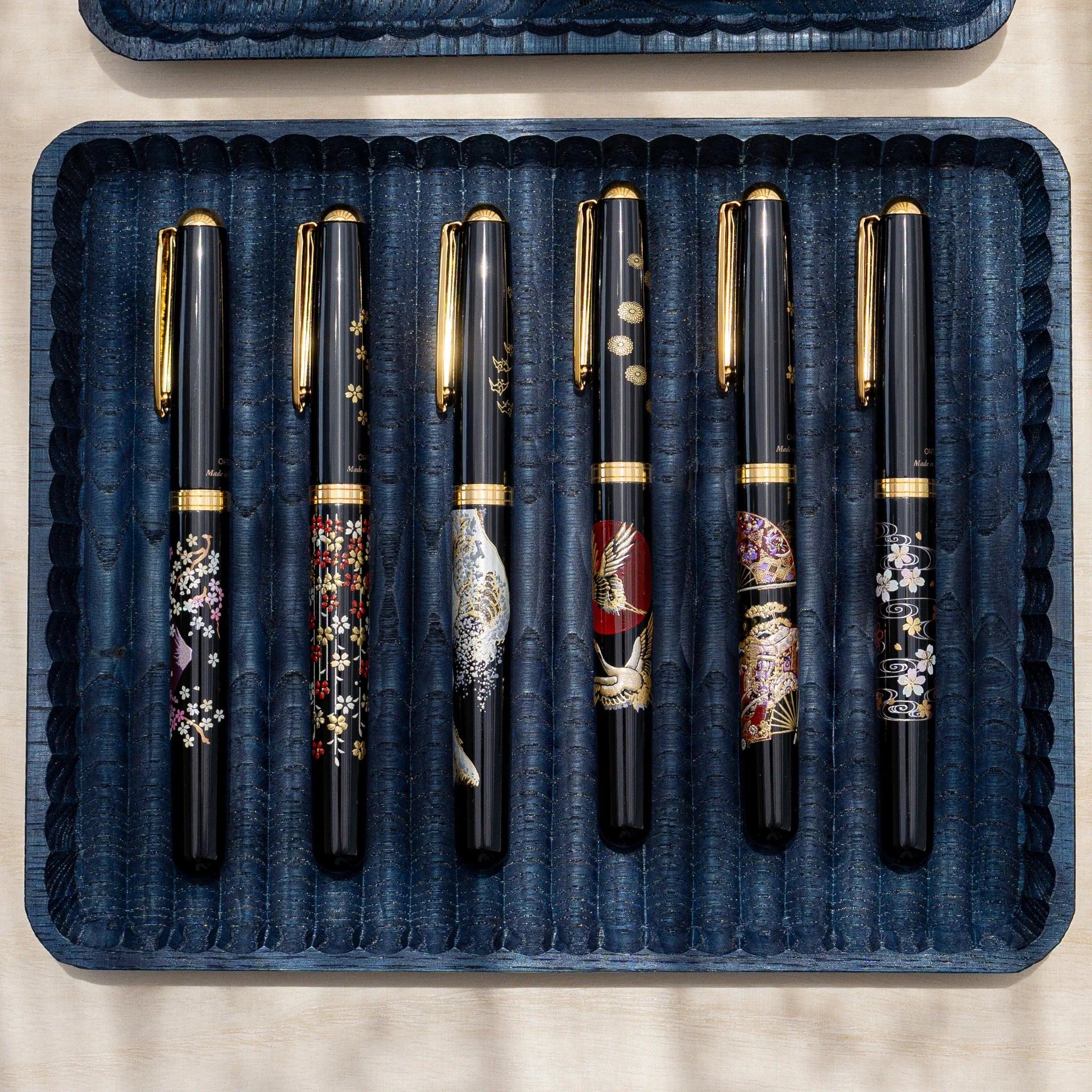 Indigo dyed wide chestnut pen tray from above holding six black fountain pens with gold and colored designs