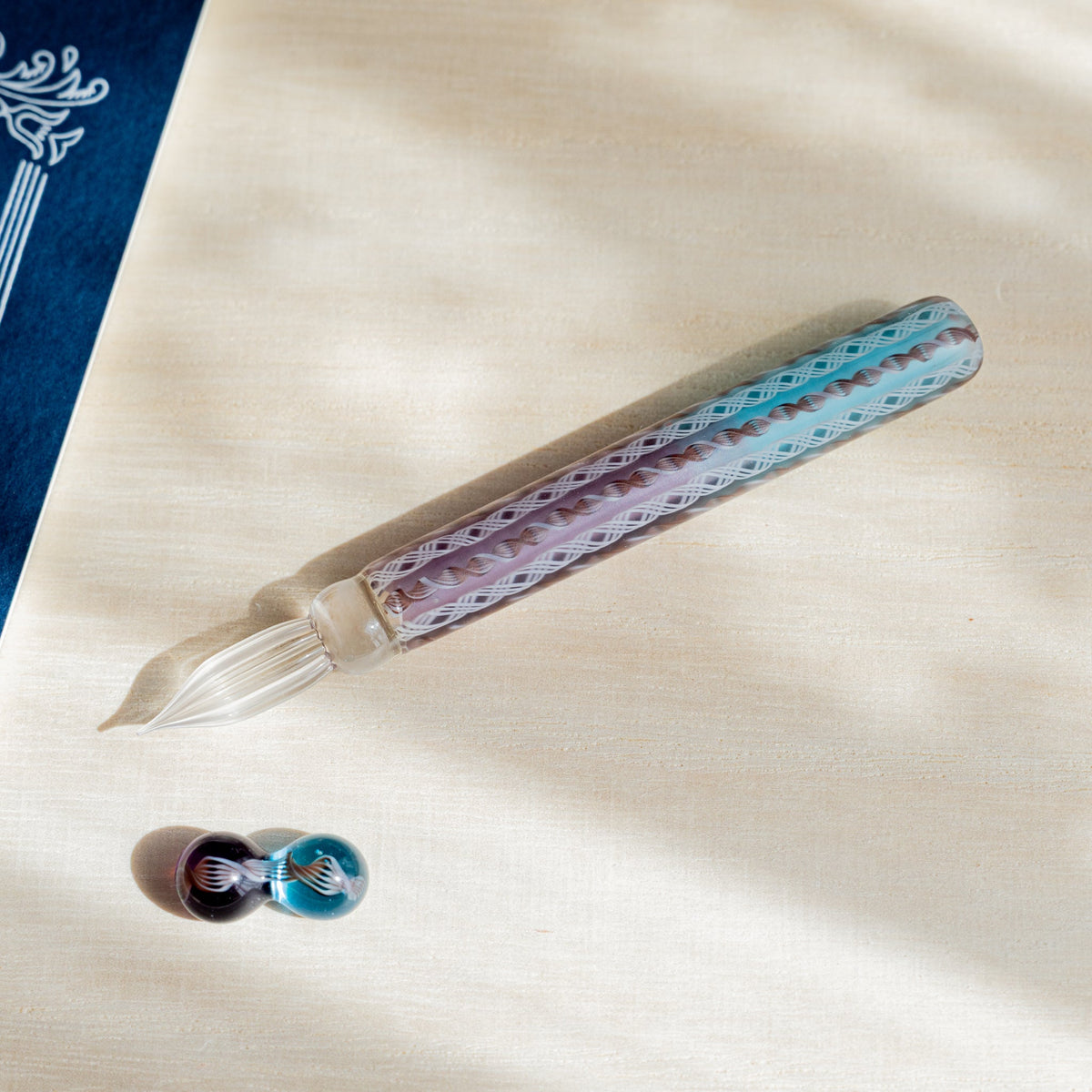 Helix Dawn glass dip pen angled across pale wood with its two-sphere glass pen rest below the nib