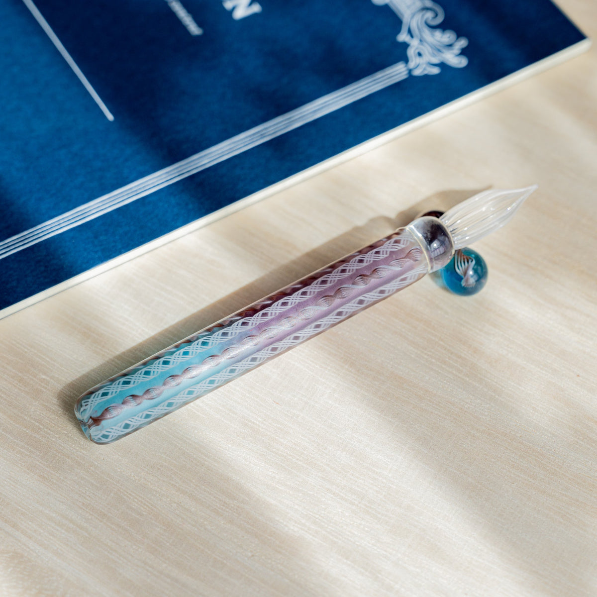 Helix Dawn glass dip pen lying horizontally on pale wood with a navy blue card and its glass pen rest