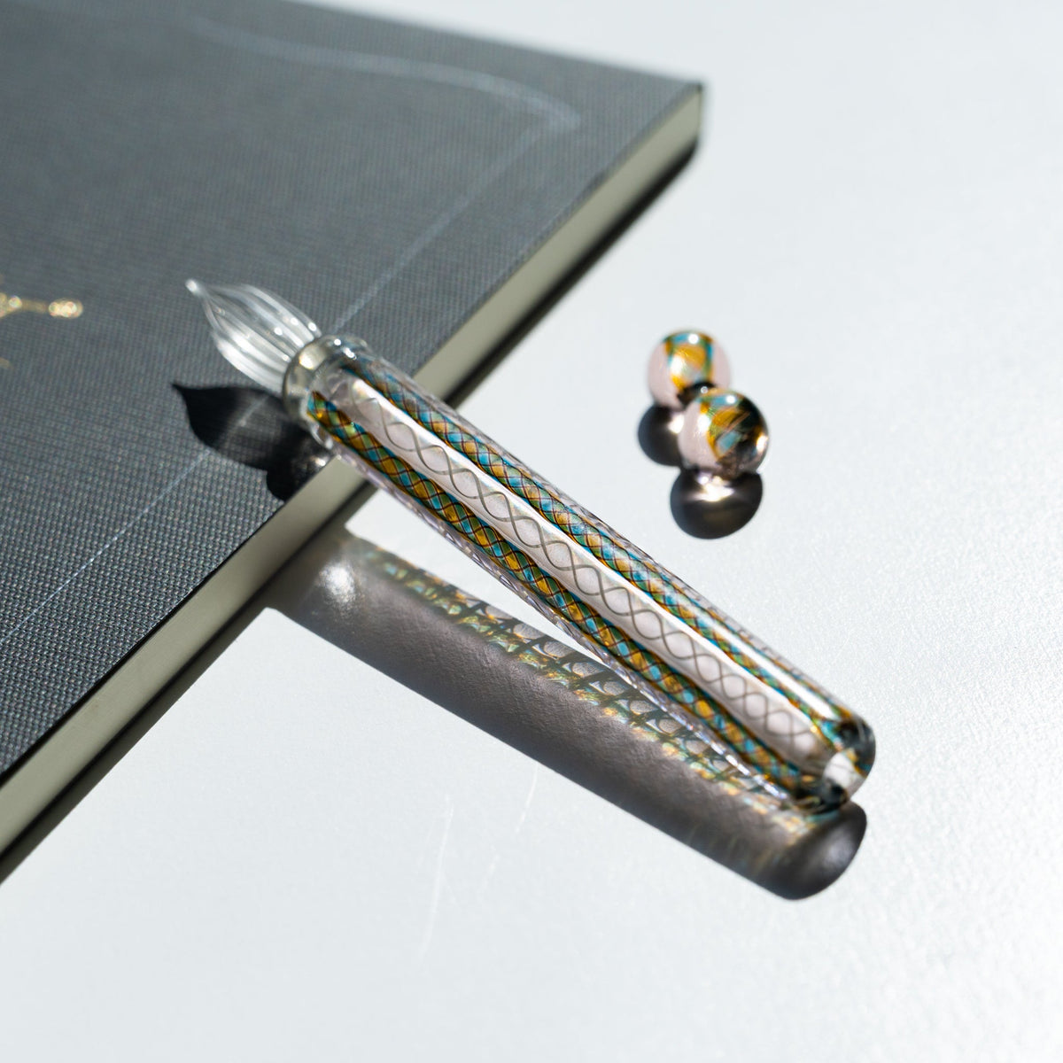 Helix Peacock glass dip pen angled off a charcoal notebook edge, glass pen rest set to one side