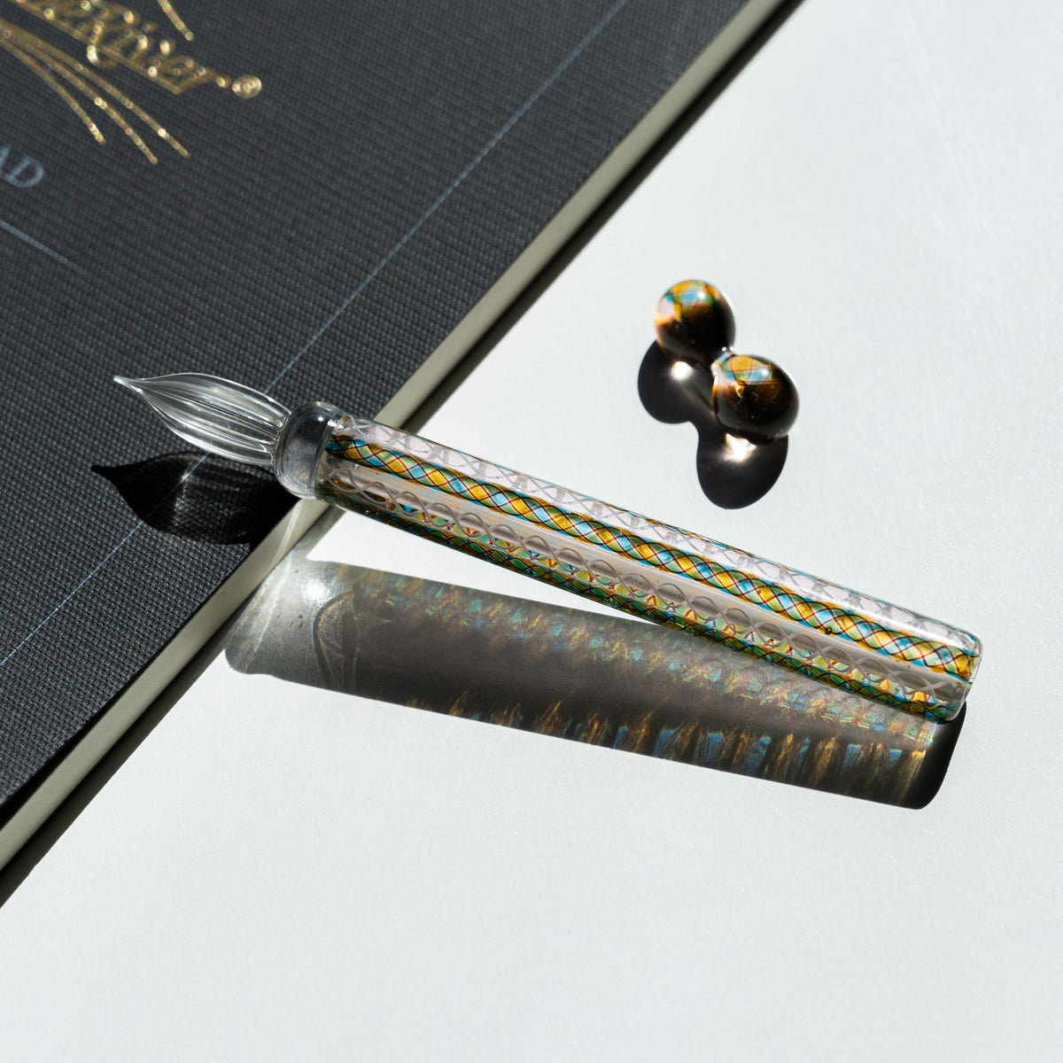 Helix Peacock glass dip pen laid on the corner of a charcoal notebook with gold foil printing