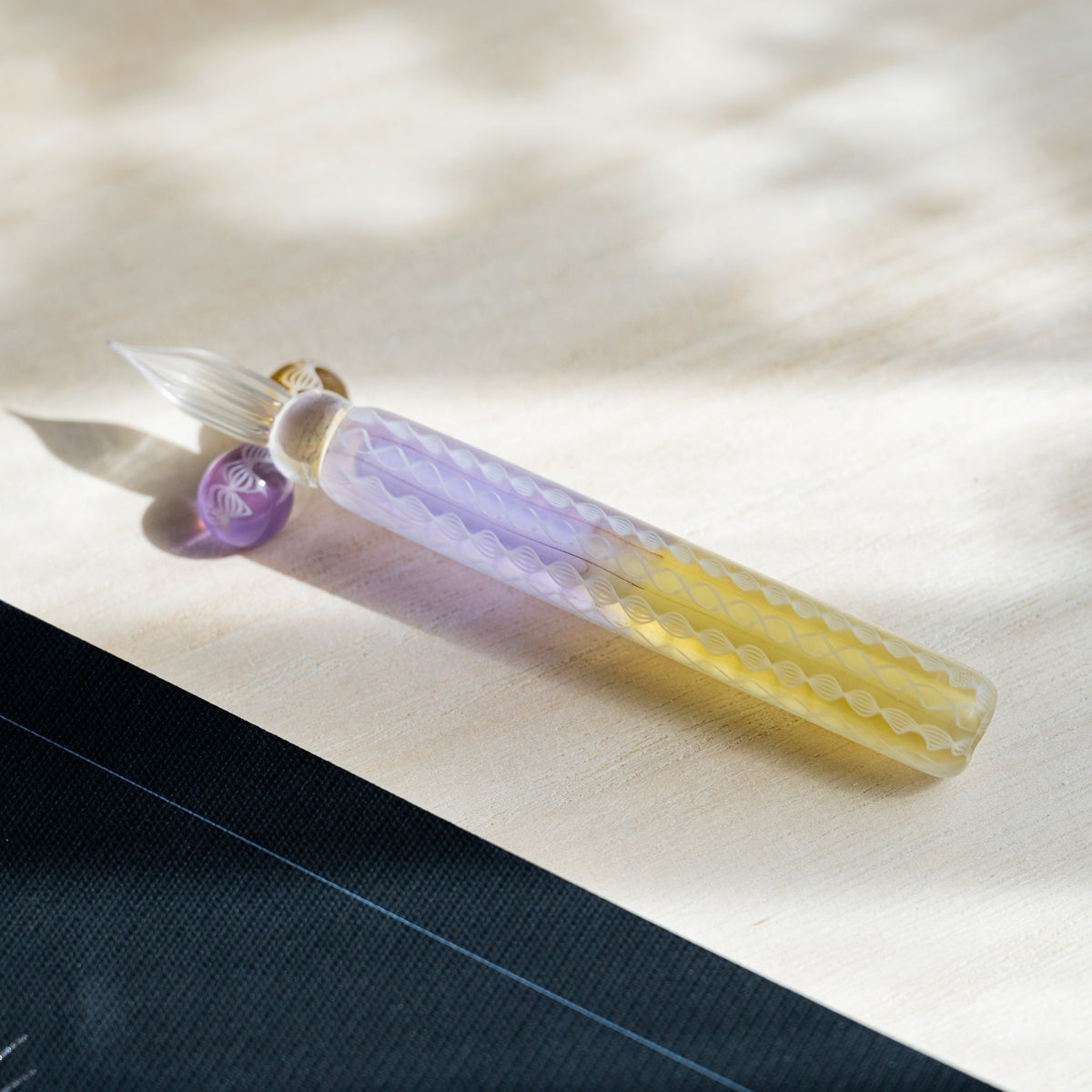 Helix Wisteria glass dip pen angled on pale wood, clear fluted nib resting on the lavender glass sphere