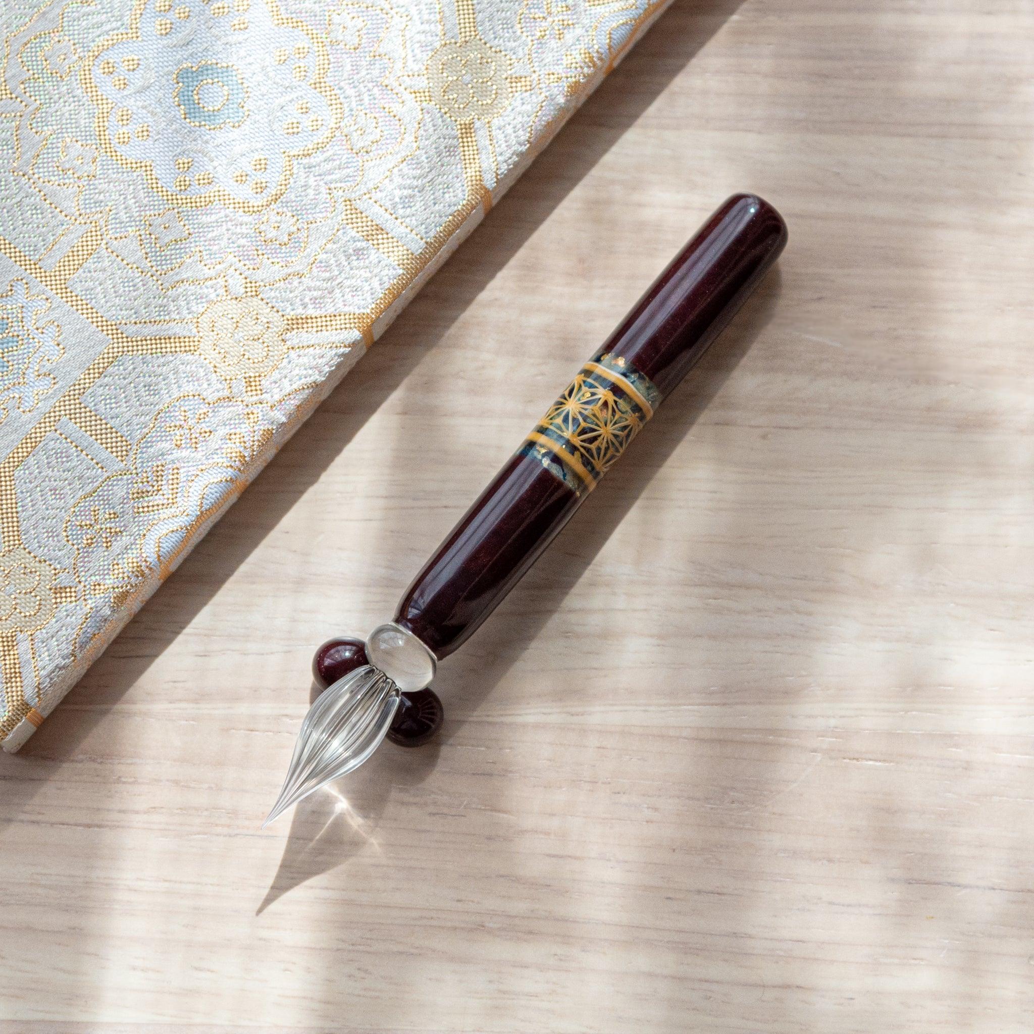 Dark brown glass pen featuring a traditional Hemp Leaves pattern in gold, paired with a matching glass pen rest. Displayed on a wooden surface with a Nishijin textile notebook.