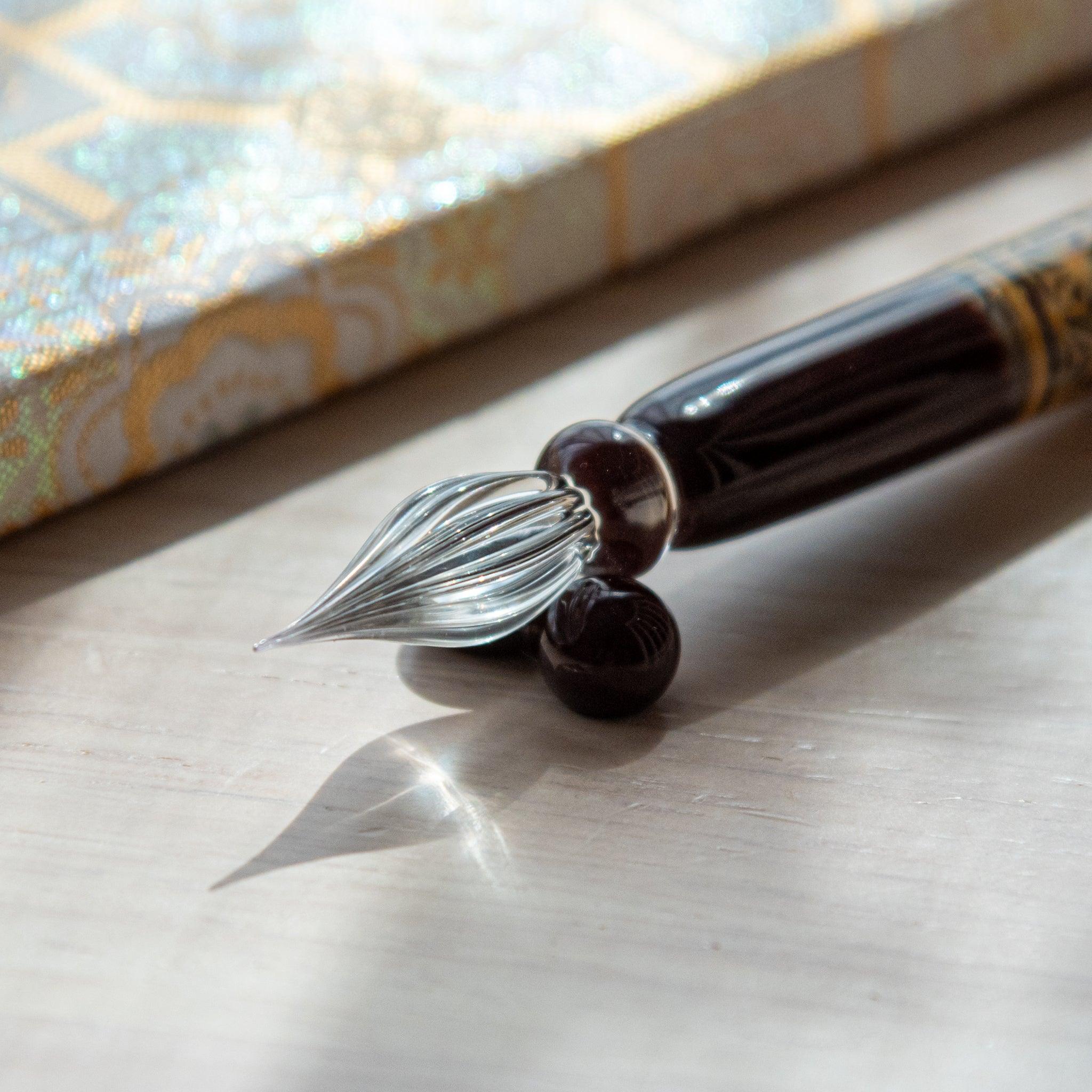 Dark brown glass pen featuring a traditional Hemp Leaves pattern in gold, paired with a matching glass pen rest. Displayed on a wooden surface with a Nishijin textile notebook.