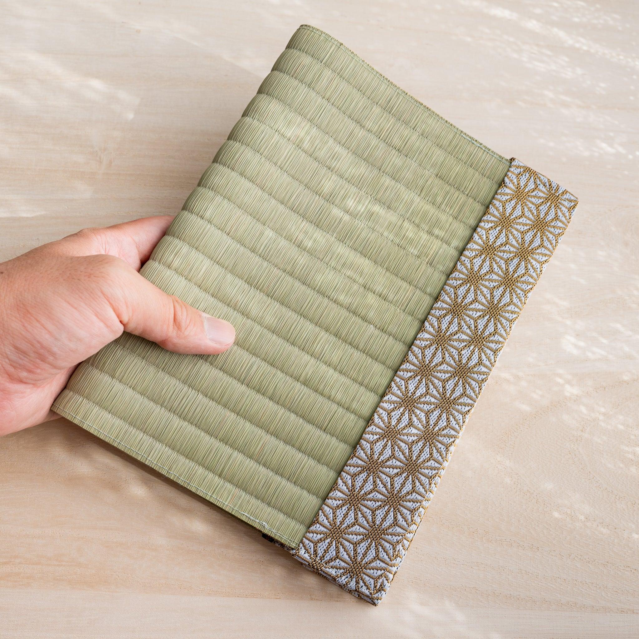 Hand holding closed tatami book cover, green rush straw front with gold and white Japanese hemp leaf fabric band
