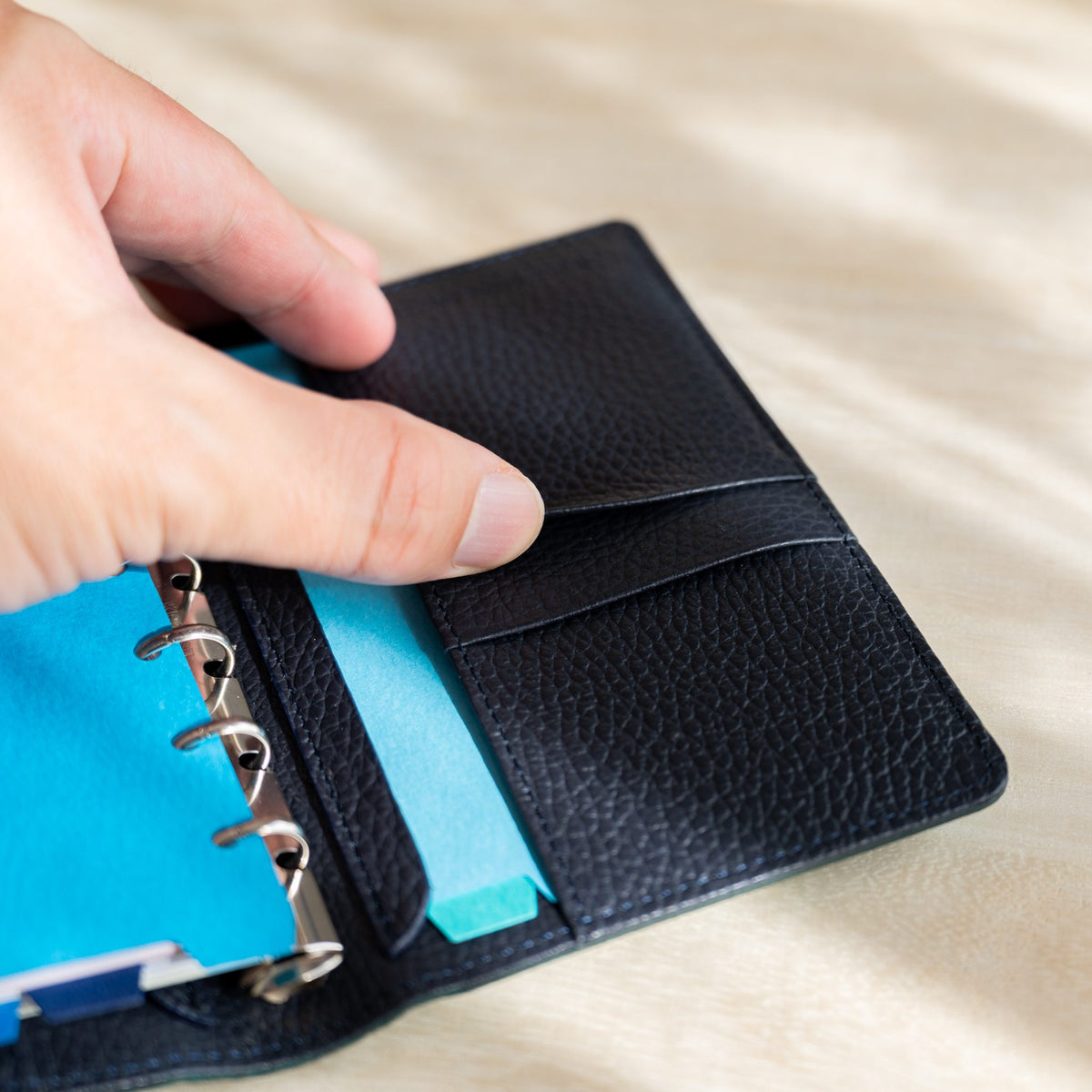 Hand opening the card pockets of the navy planner, blue tabbed pages on the rings at left