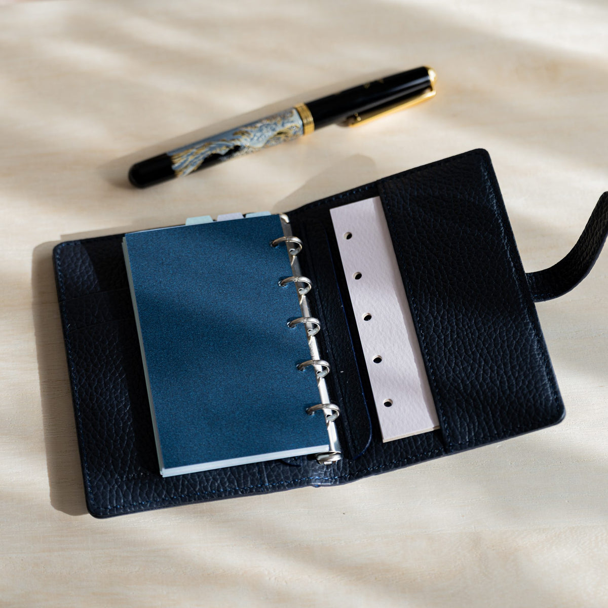 Open navy planner with a dark blue sheet on the rings, pale pink punched strip and fountain pen above