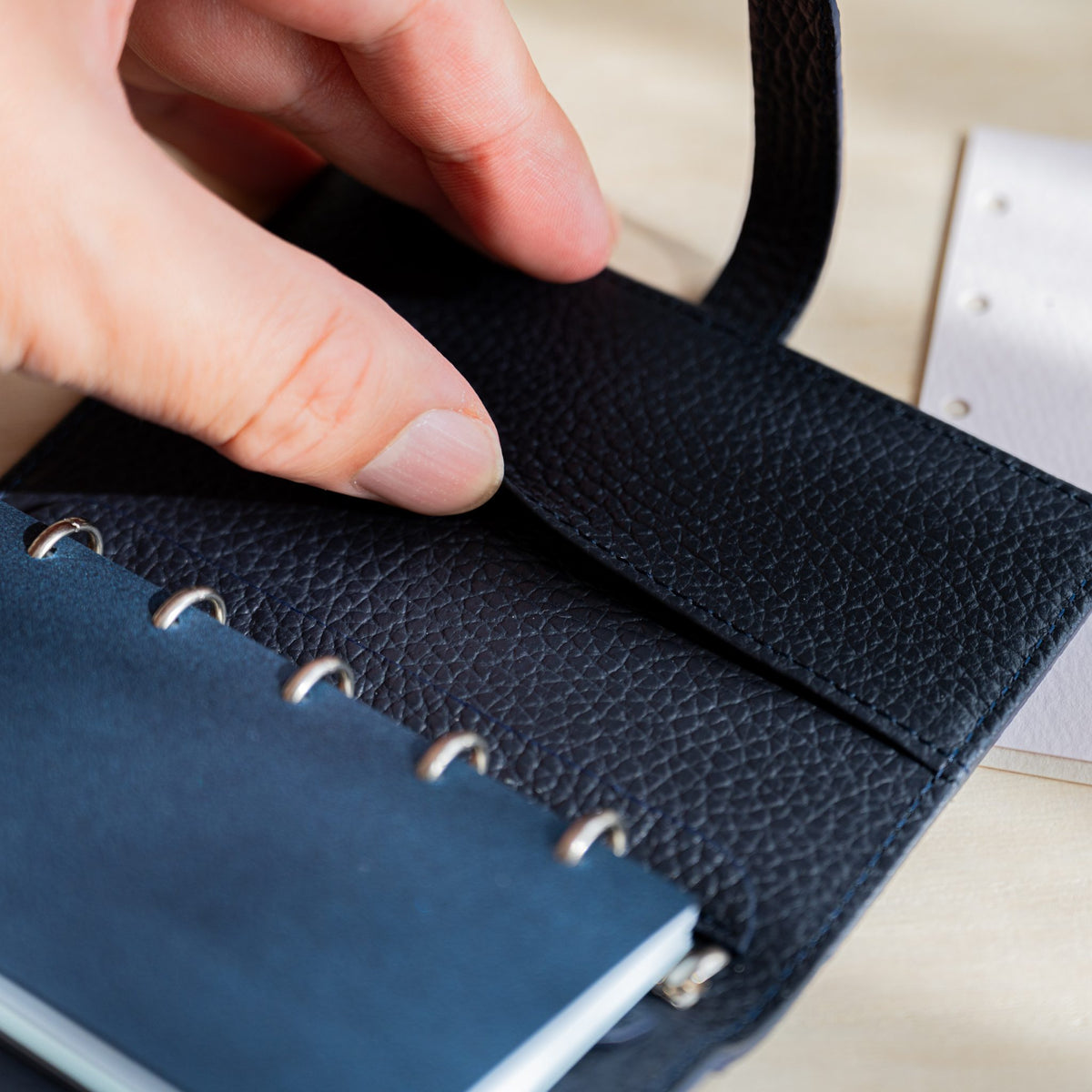 Hand lifting the pocket flap of the navy planner above a dark blue sheet on the rings