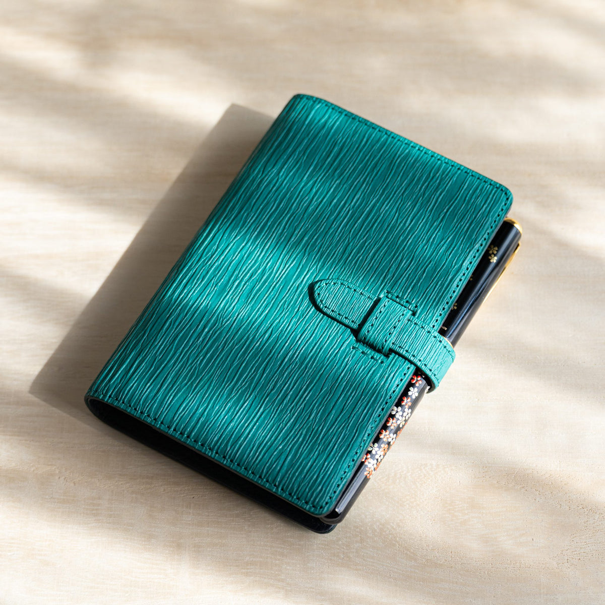 Teal Ise Gikakushi planner with a black fountain pen with floral pattern held by the strap