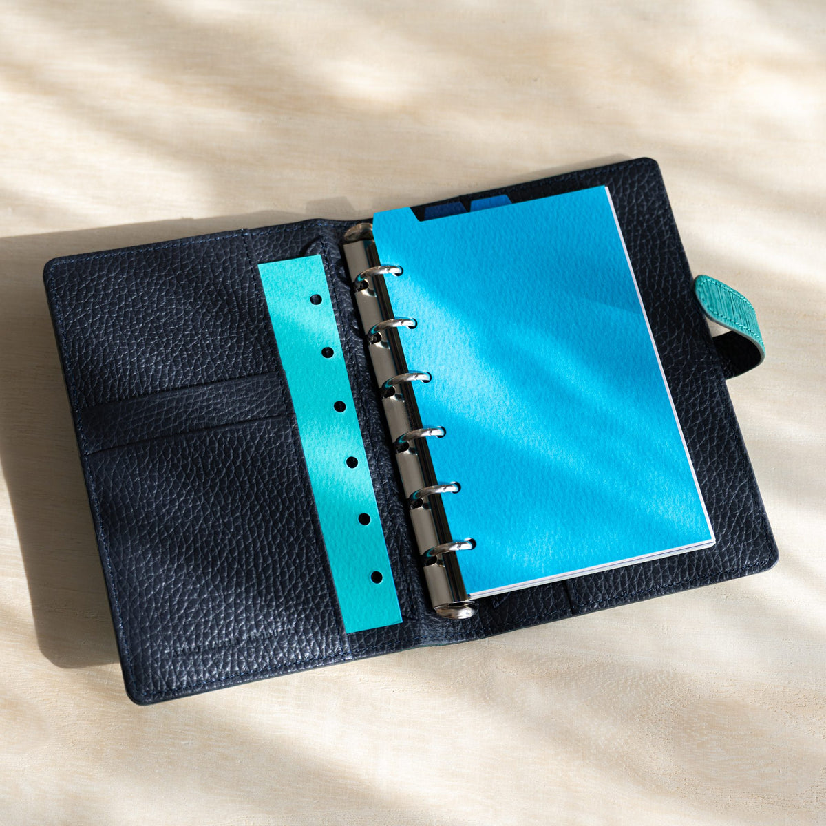 Open planner with a turquoise hole-punched strip in the pocket, sky blue divider on rings and teal strap