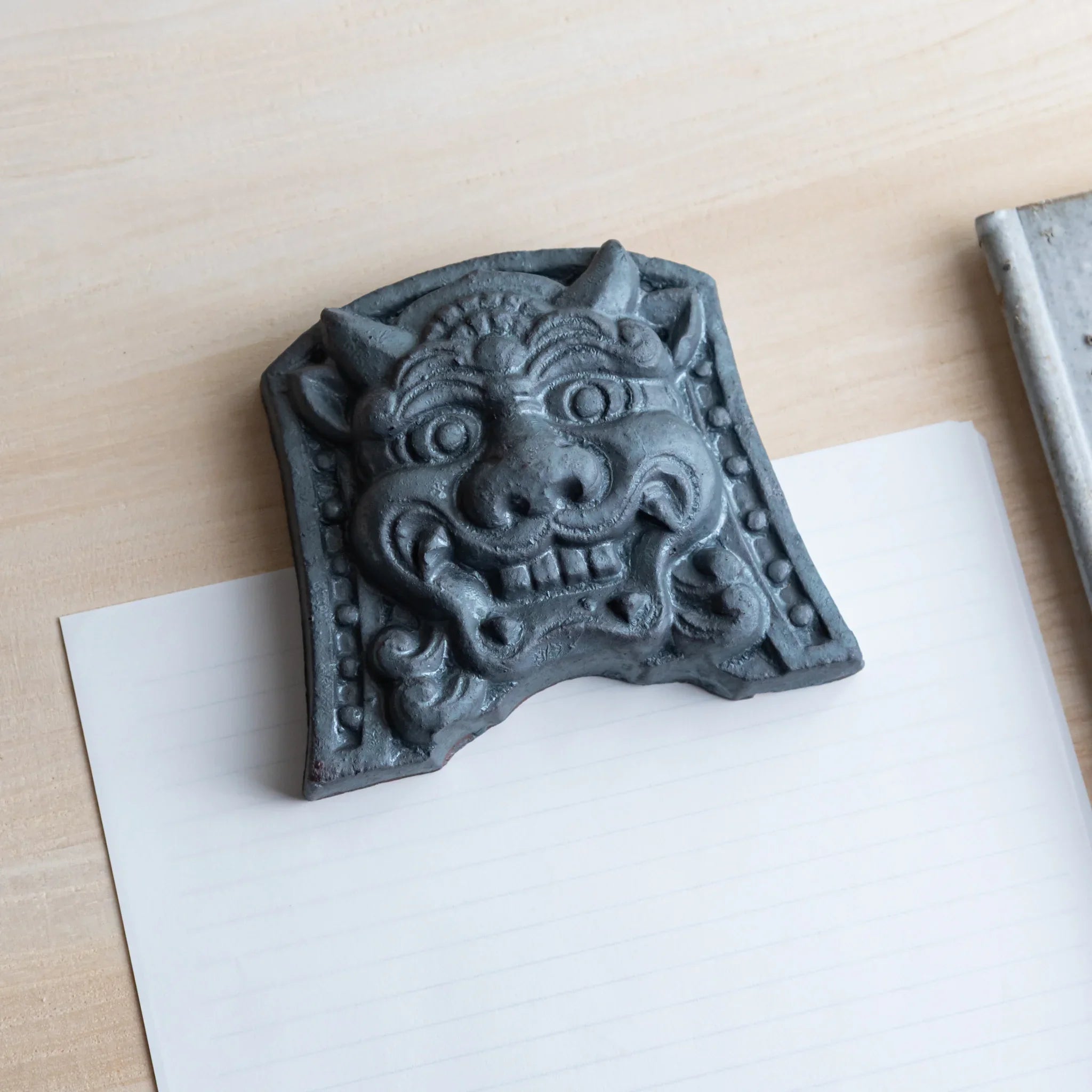 Japanese Paperweight Desk Accessory featuring resolute oni face ceramic tile angled on lined paper with rod.