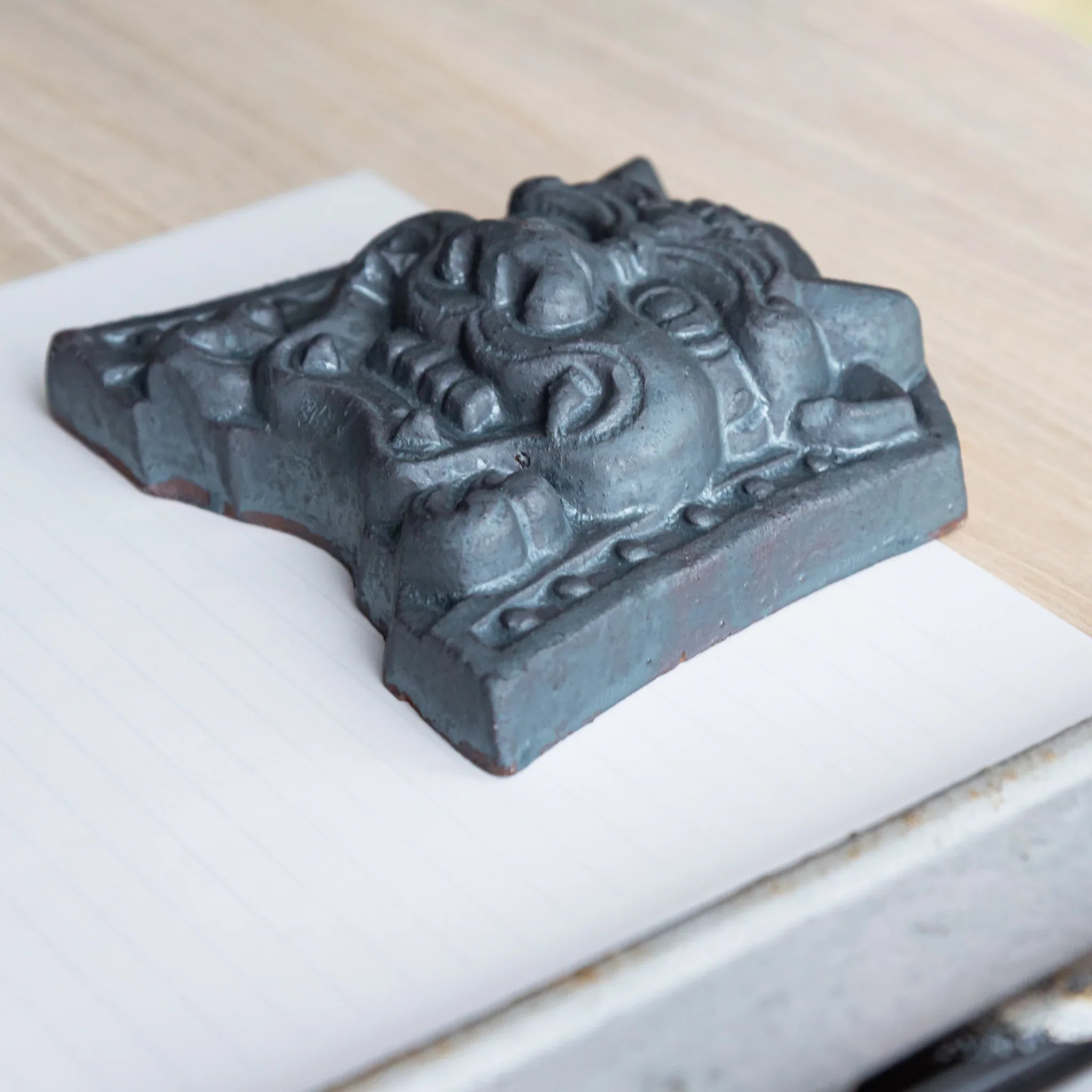 Japanese Paperweight Desk Accessory depicting resolute oni face ceramic tile lying on lined paper viewed from side.
