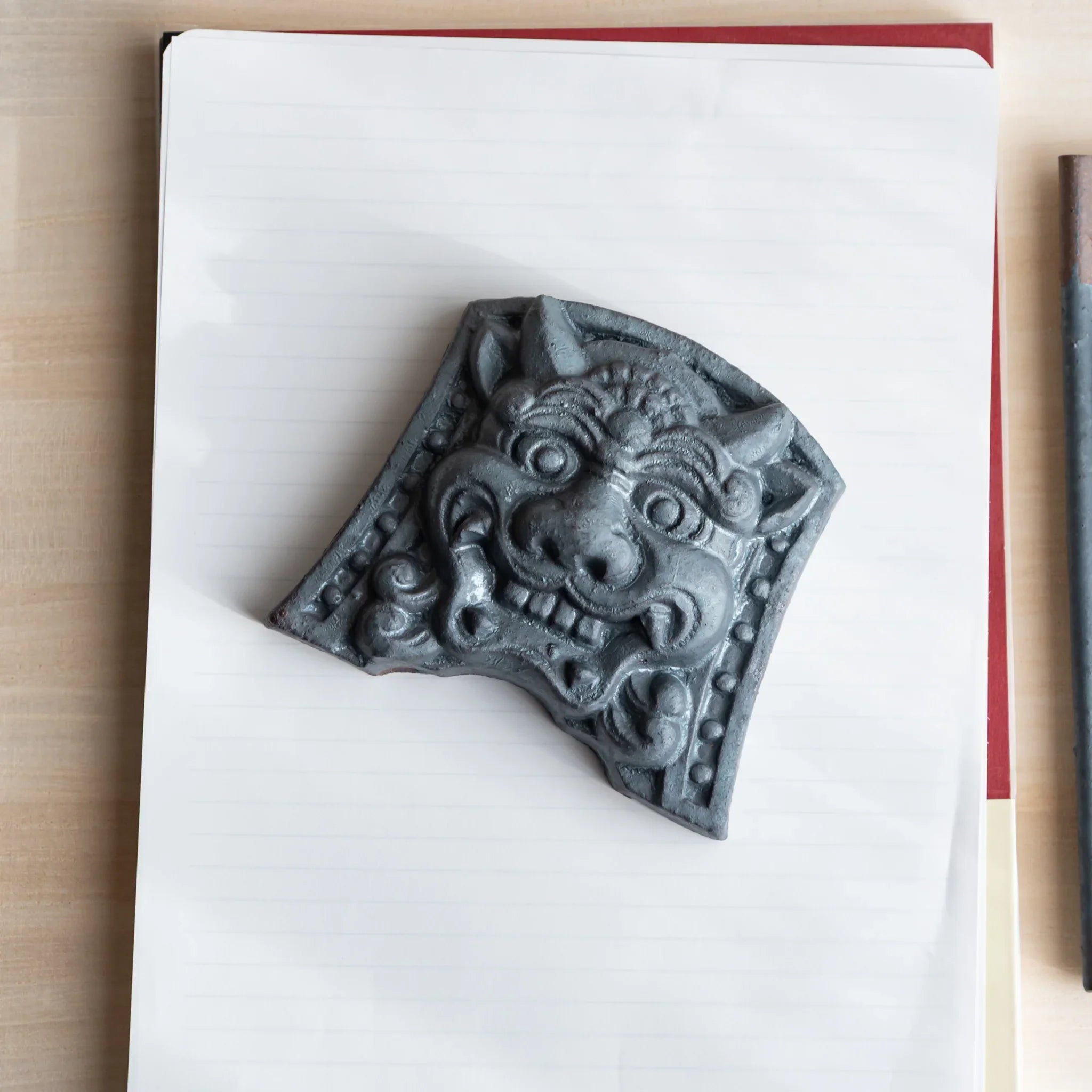 Japanese Paperweight Desk Accessory depicting resolute oni roof guardian tile angled on lined paper notebook.