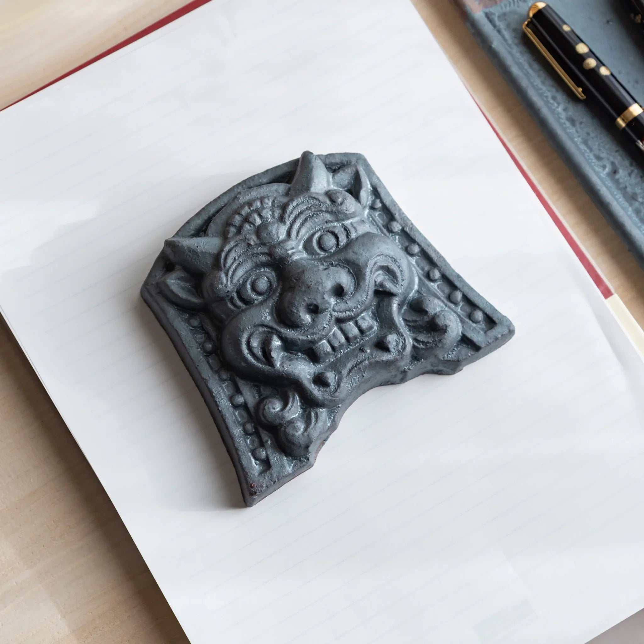 Japanese Paperweight Desk Accessory featuring resolute oni roof guardian tile on lined paper with pen tray and pen.