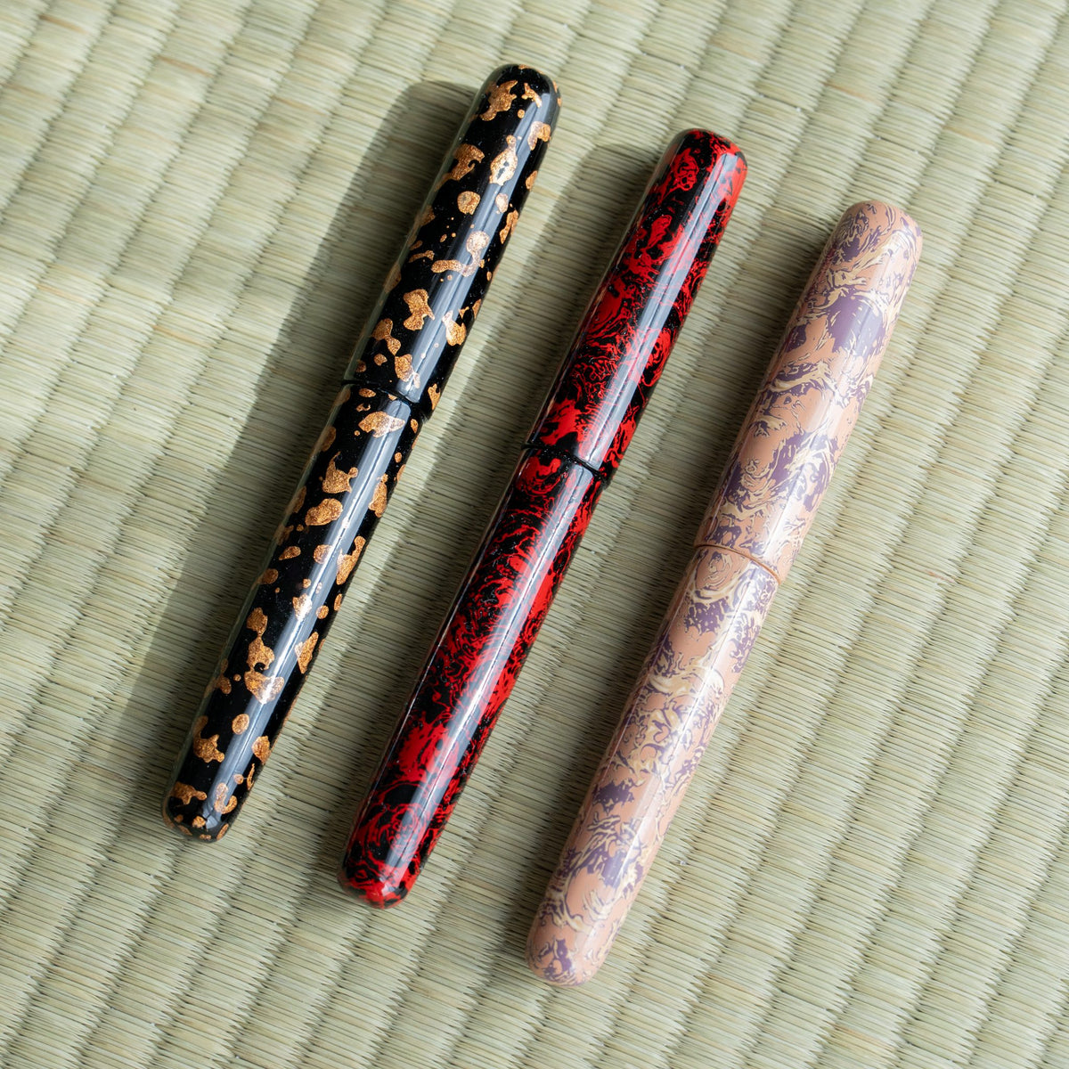 Three Tsugaru-nuri urushi fountain pens laid diagonally on tatami: black with gold flecks, red and black, and pink with purple pattern