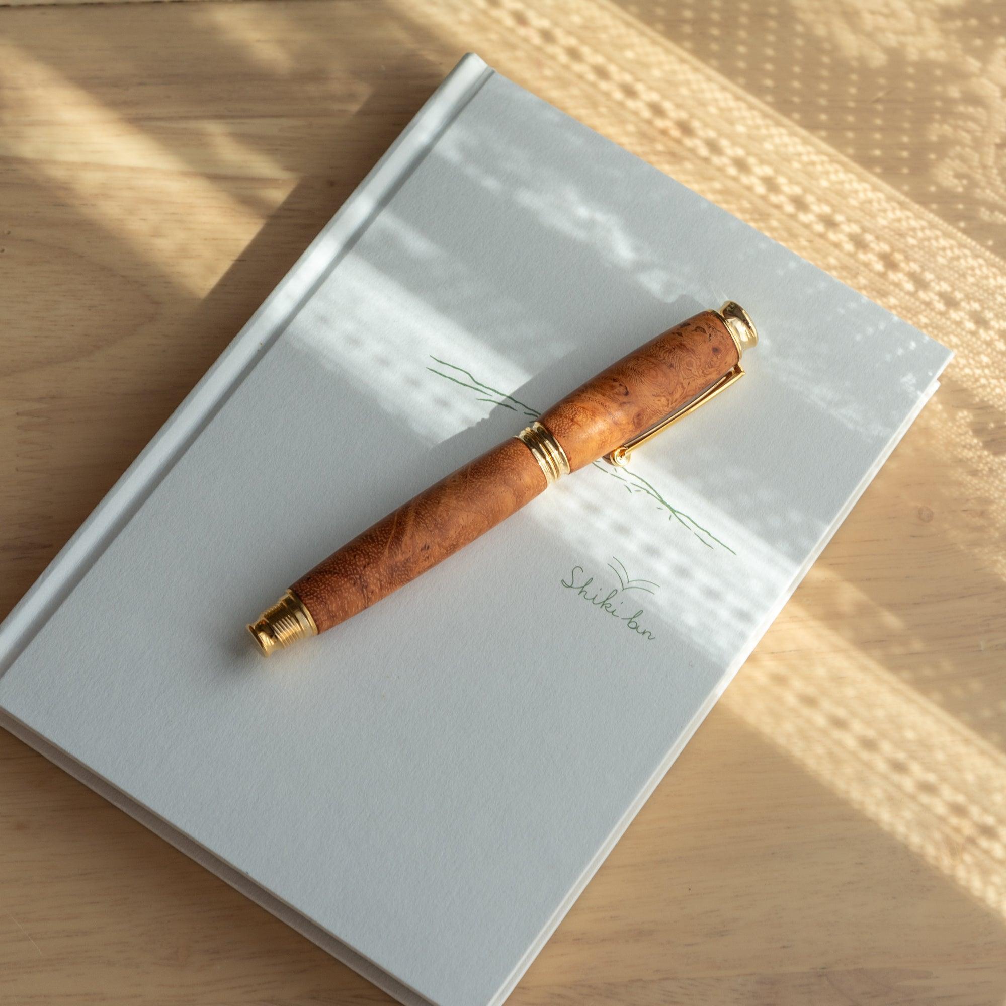 A Japanese fountain pen with a Karin wood barrel and gold accents, resting on a white notebook under soft light patterns, showcasing its elegance.