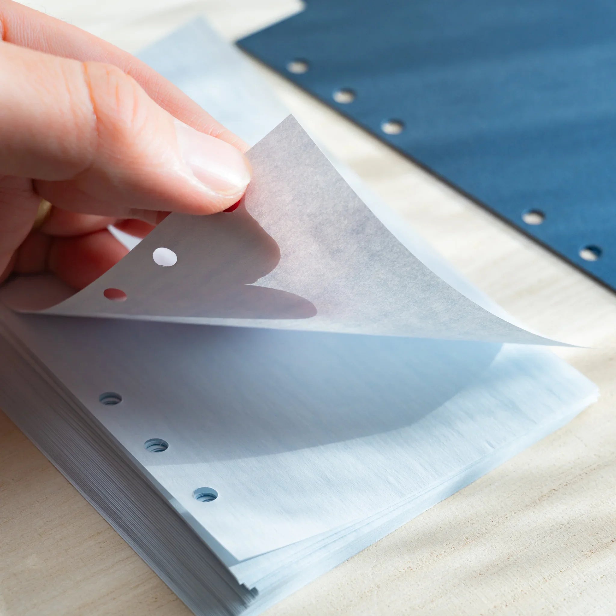 Hand lifting a thin pale blue onion skin planner sheet, showing its translucency and six-hole punches. 