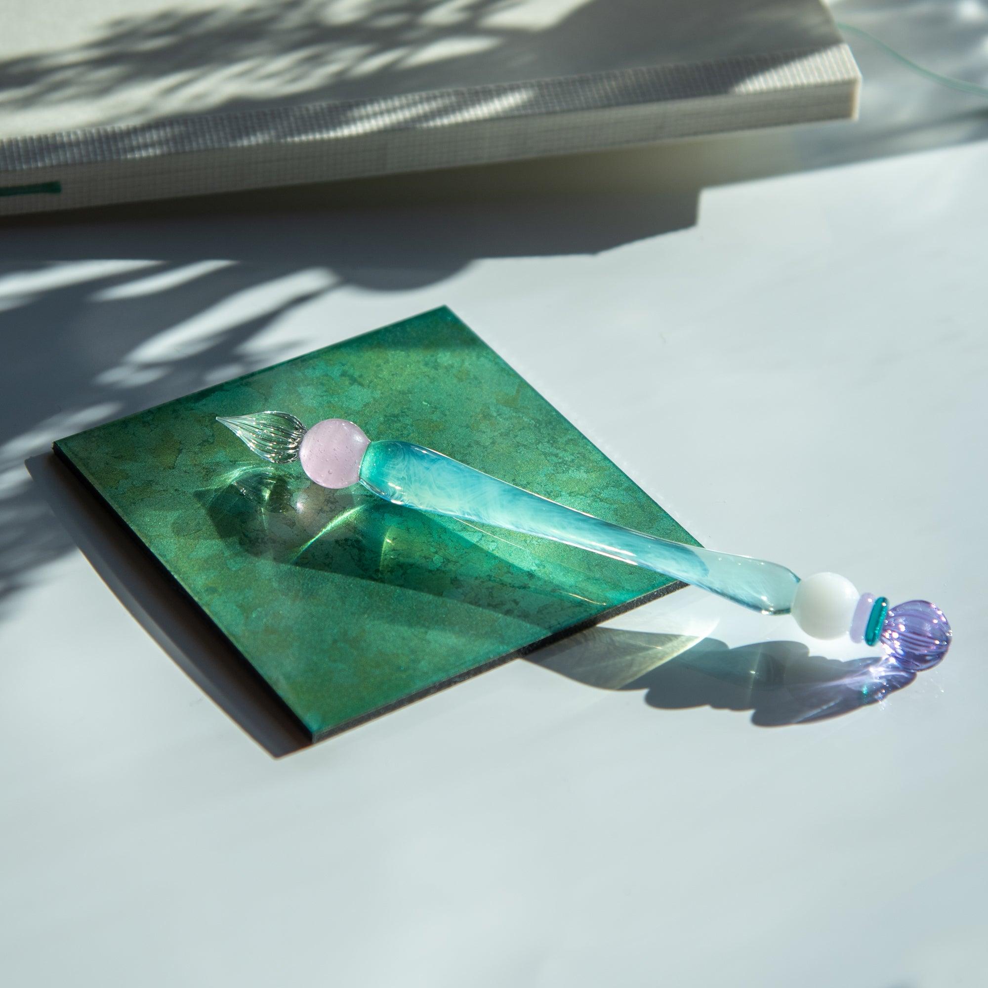 A pastel-themed glass pen with a purple candy-like ornament and blue body, resting on a green tile with soft sunlight illuminating it.