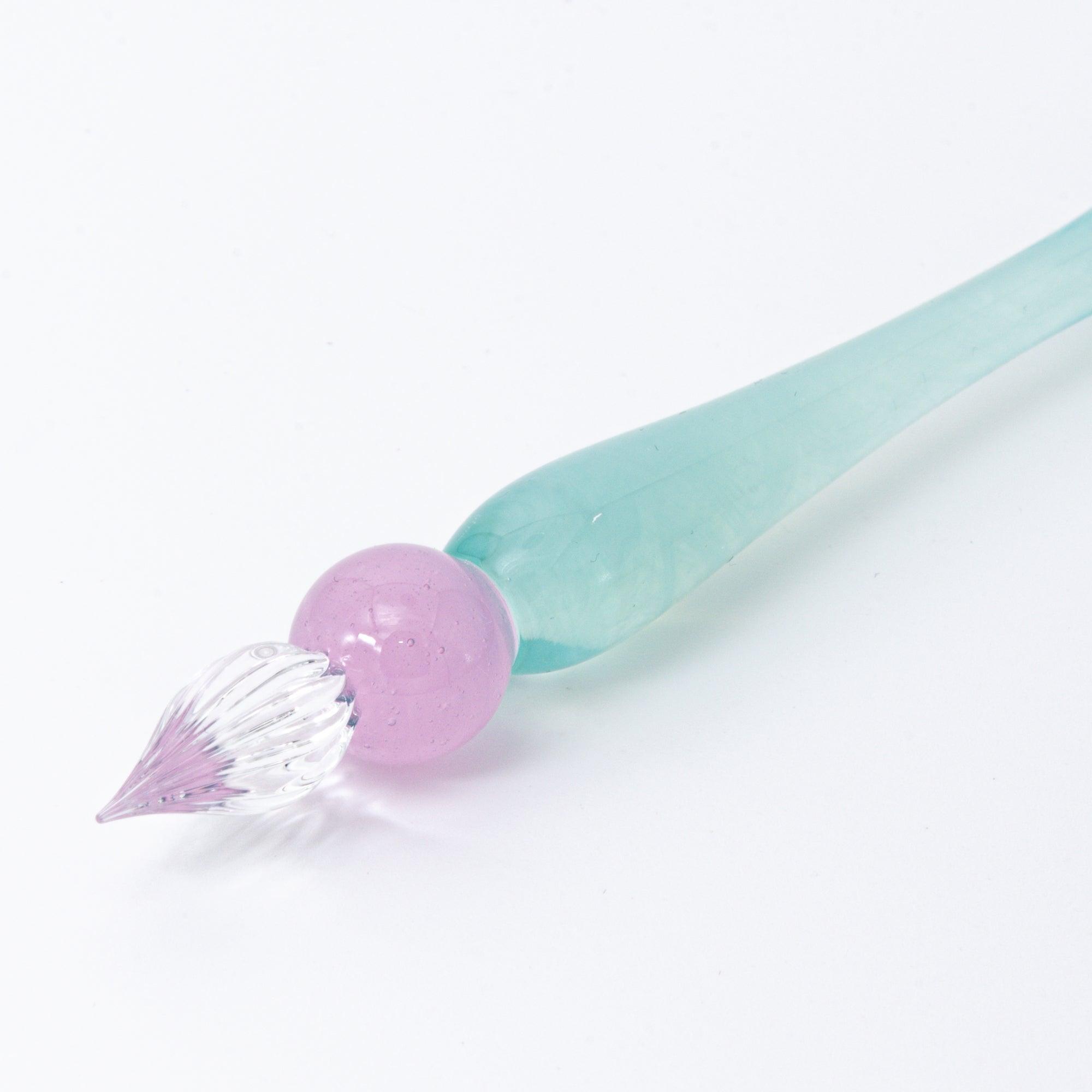 A pastel-colored glass pen with a soft blue body and candy-like top rests on a notebook, bathed in soft sunlight.