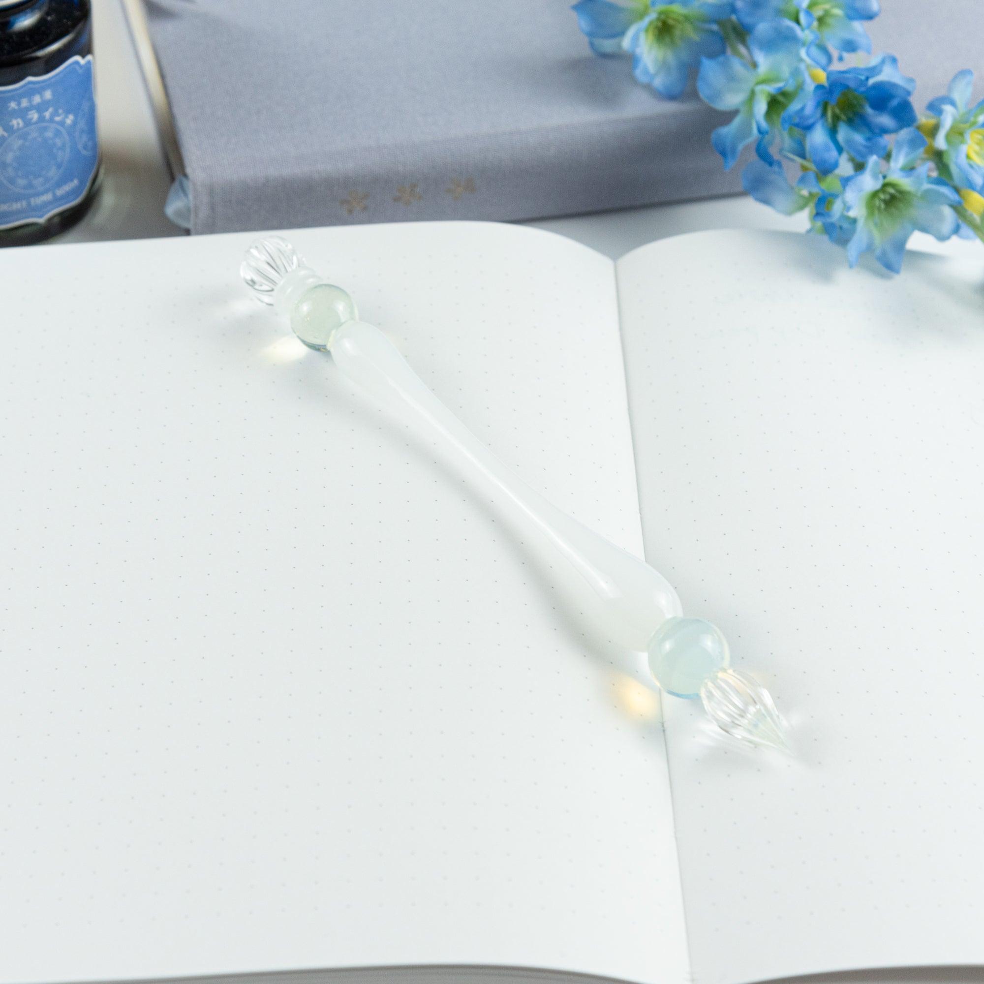 White glass pen with light blue spheres laid across an open notebook on a desk, inviting creativity and elegant writing.