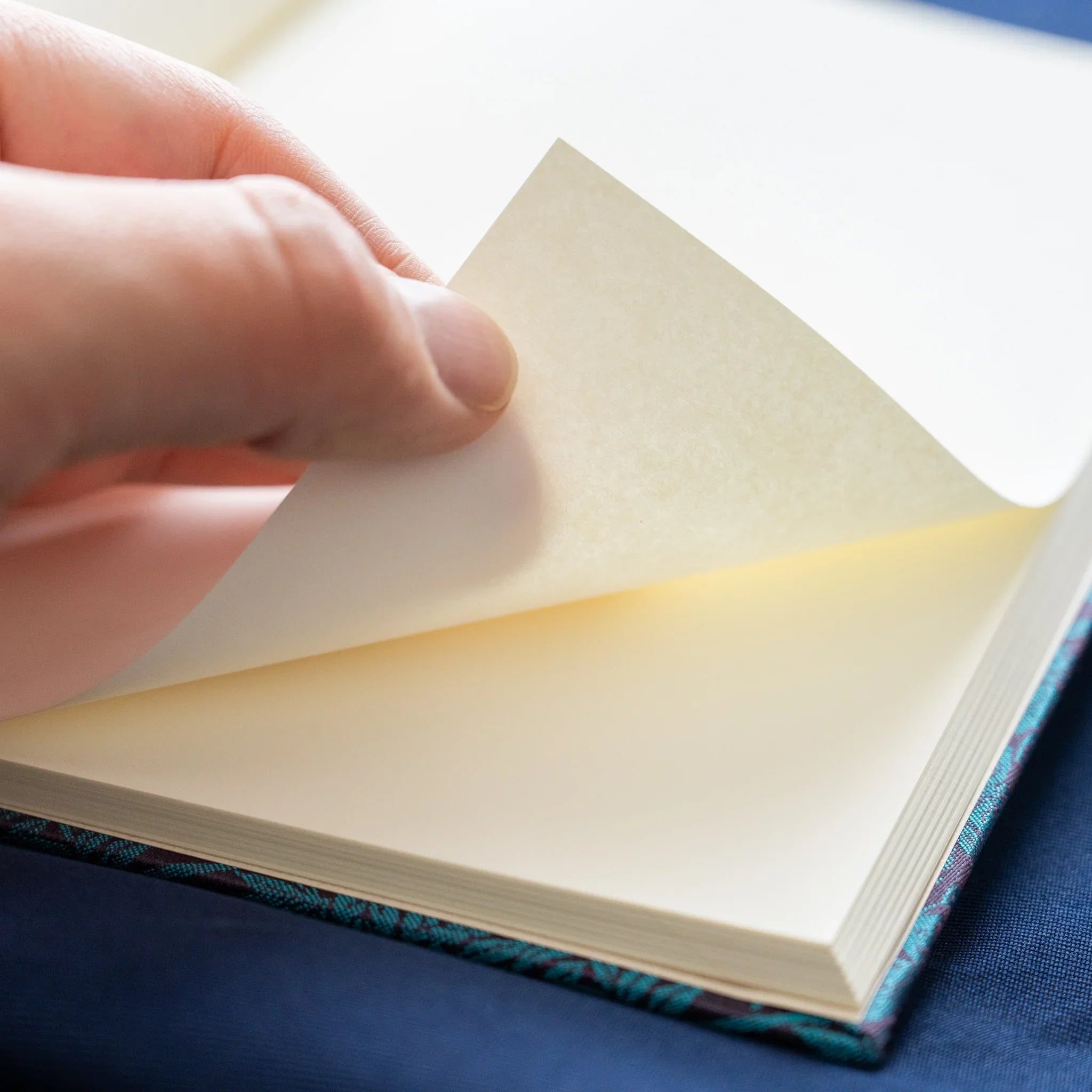 Hand turning pages of Kiri Take Ho-o Nishijin silk Homeicho notebook revealing cream paper with teal cover at base
