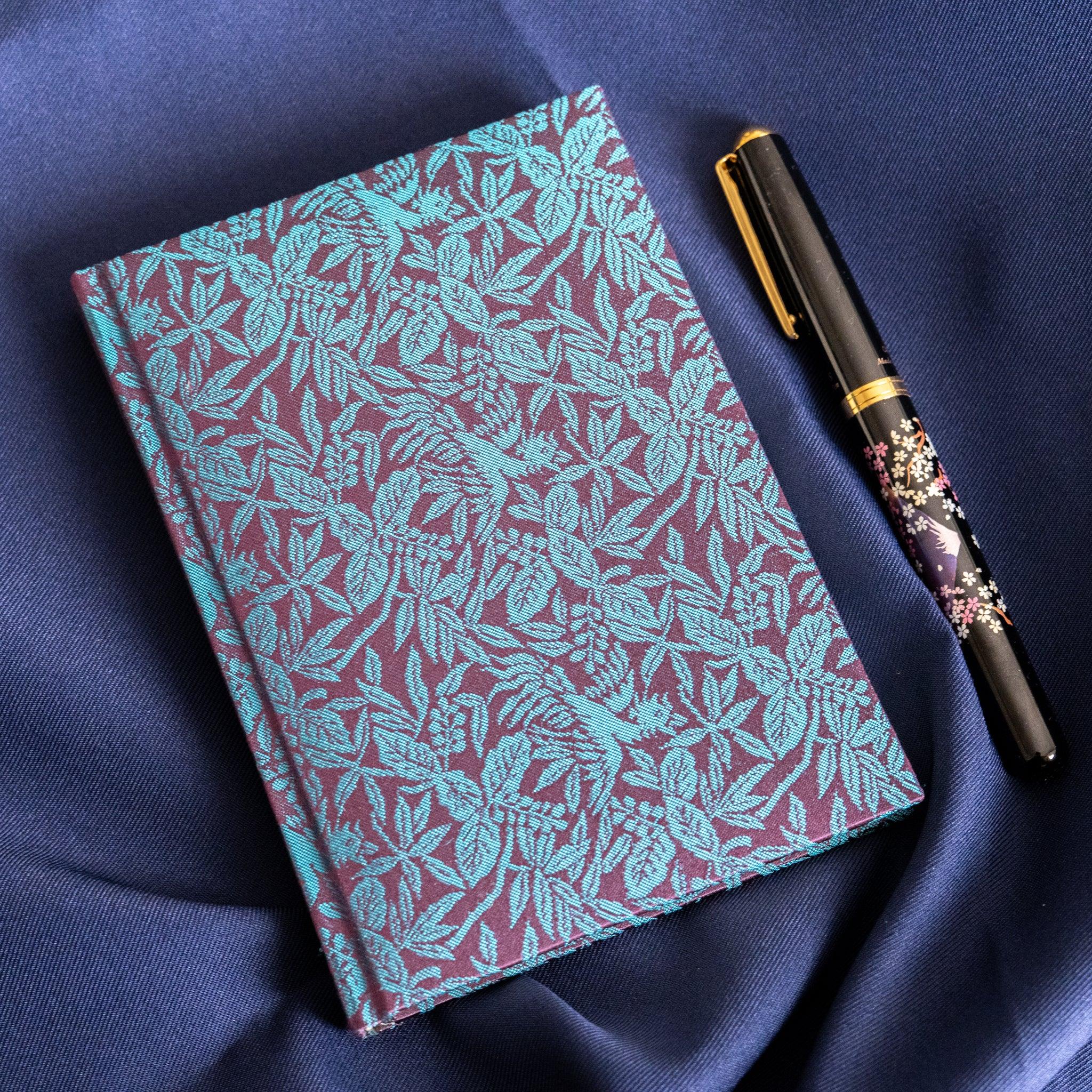 Kiri Take Ho-o Nishijin silk Homeicho notebook and decorative maki-e pen on navy fabric, angled lifestyle view
