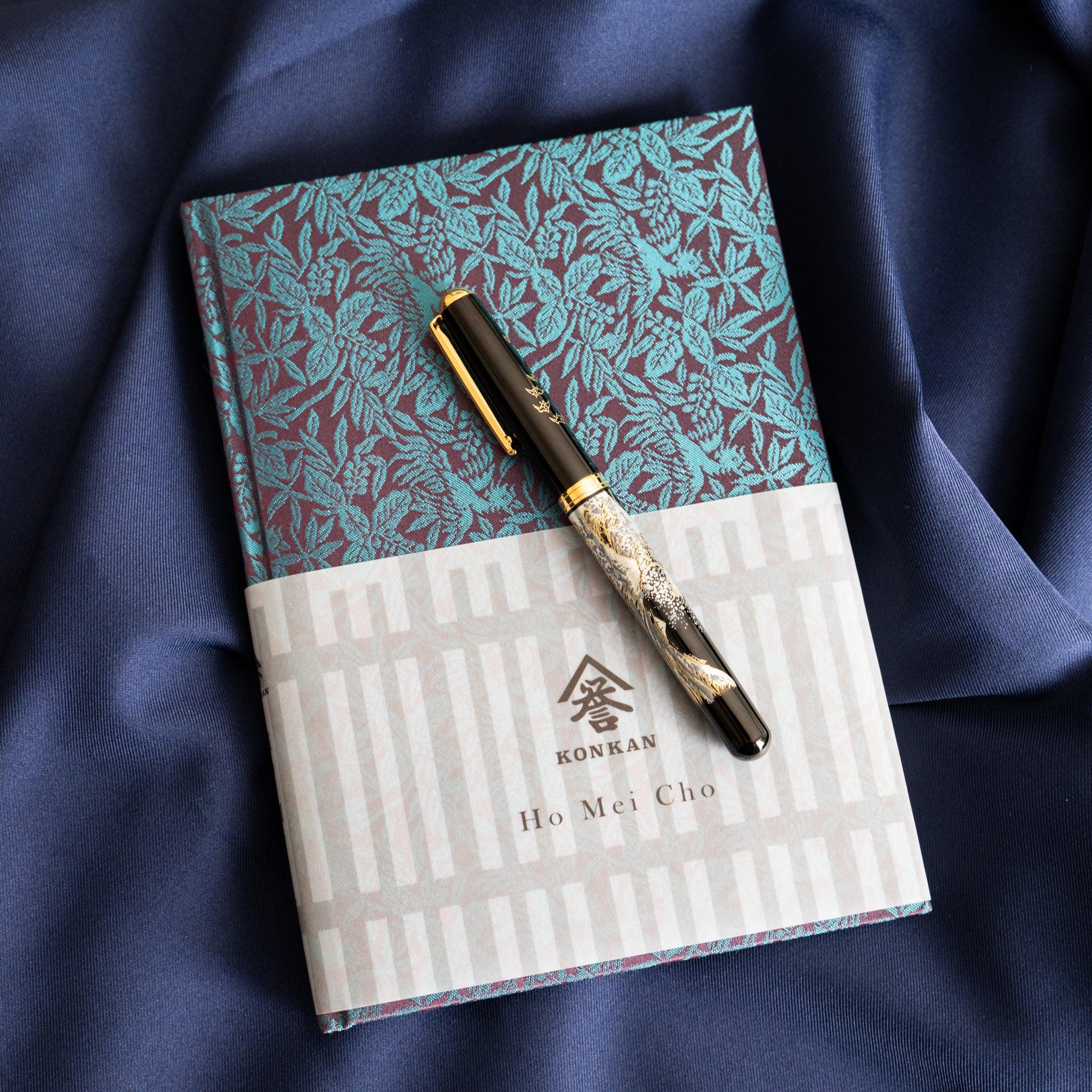 Kiri Take Ho-o Nishijin silk Homeicho notebook with paper band and fountain pen on blue fabric, product lifestyle shot