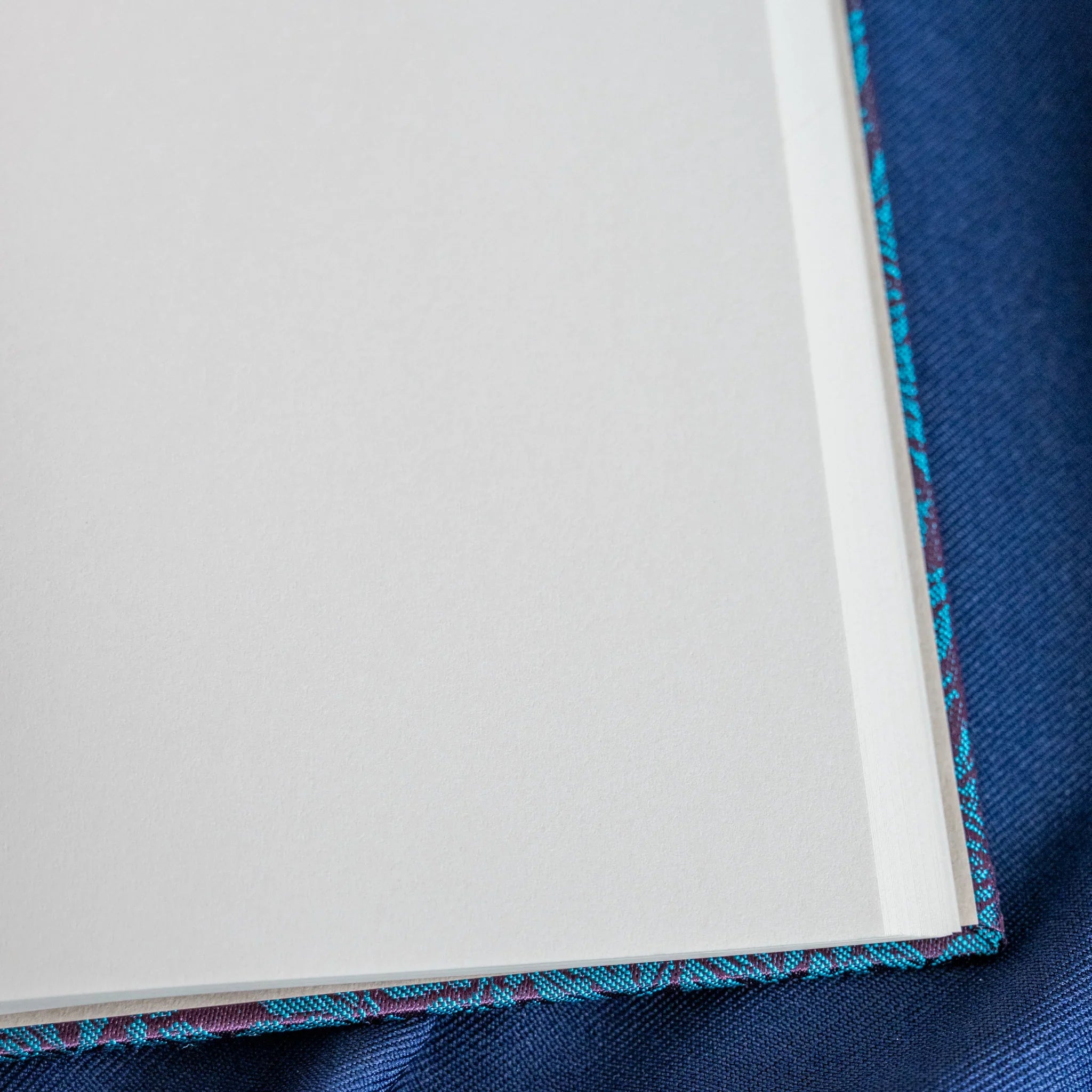 Kiri Take Ho-o Nishijin silk notebook corner showing white page stack and teal woven cover wrapping on navy fabric