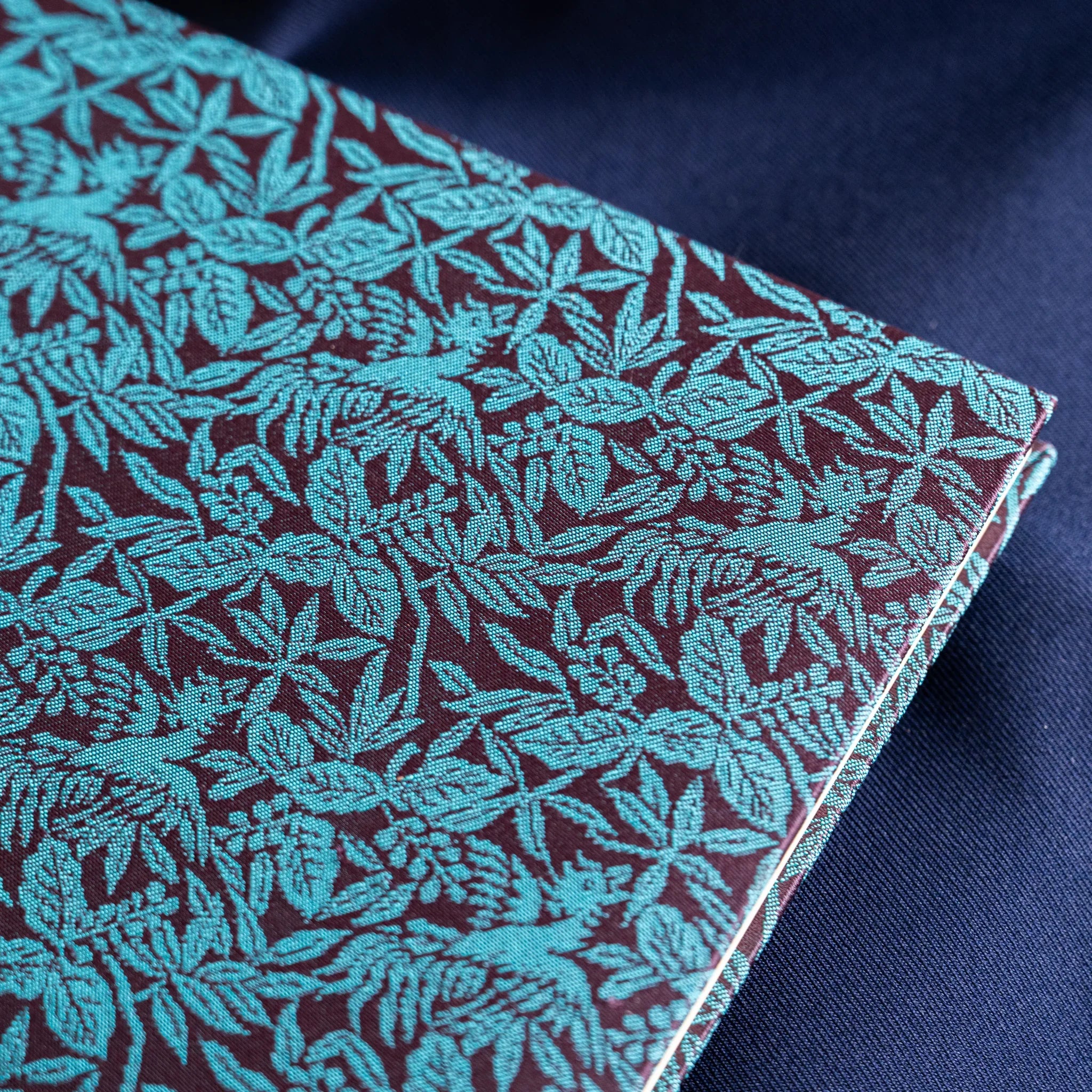 Close-up angled view of Kiri Take Ho-o Nishijin silk notebook cover showing teal leaf and floral weave pattern detail
